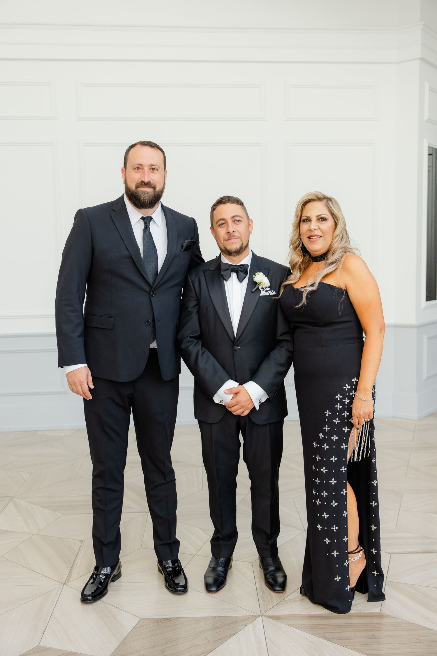 Three wedding guests pose together in formal black attire for an elegant portrait against a white backdrop.