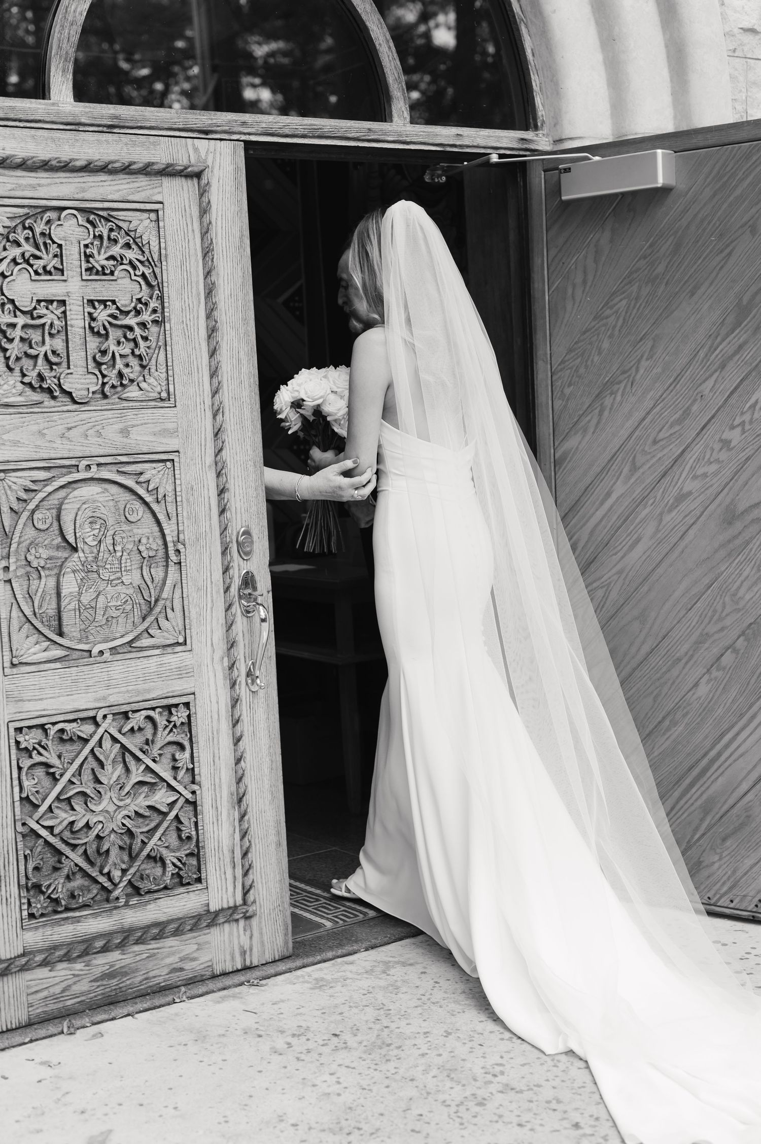 Black and white photo of bride in flowing dress and veil entering ornate church door.