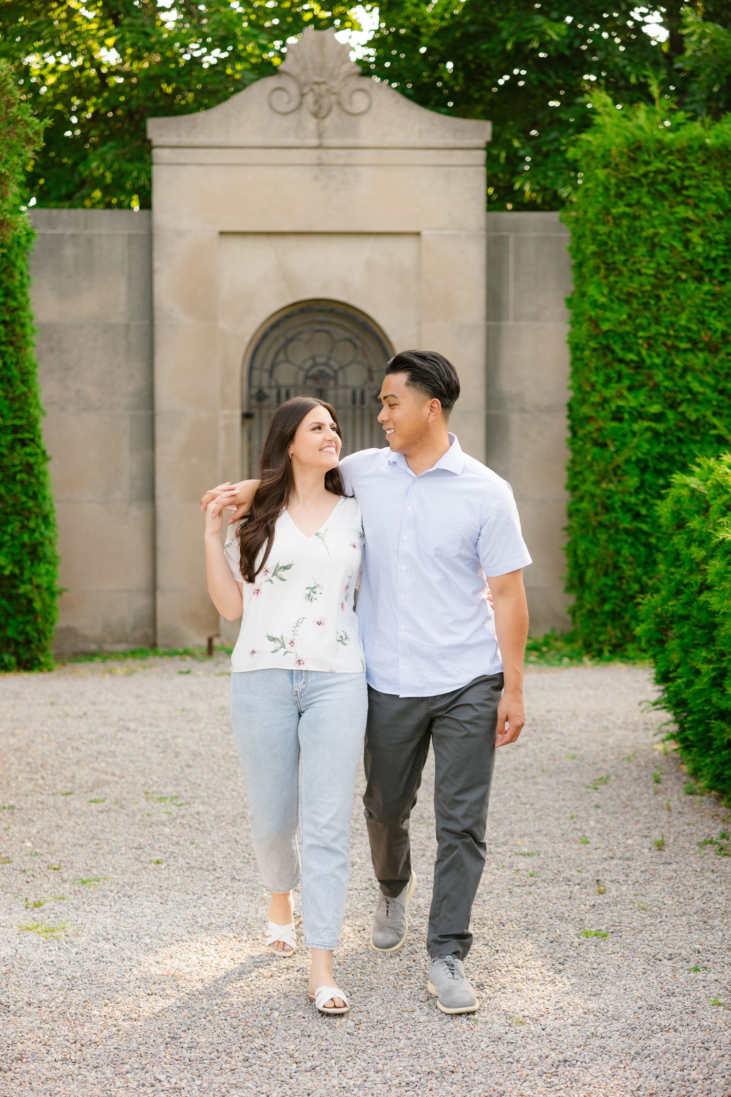 A couple walks together on a gravel path in front of an ornate stone archway surrounded by lush greenery.