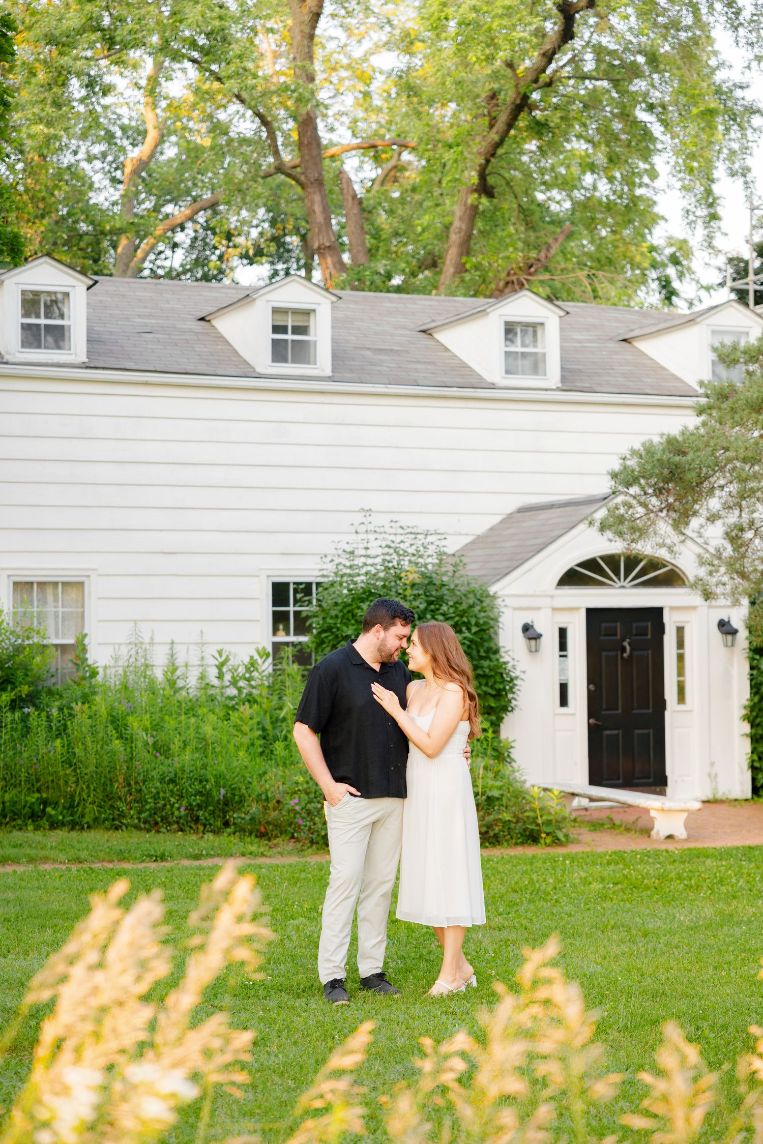 Romantic couple embracing on lawn in front of white colonial house with dormer windows and black door.