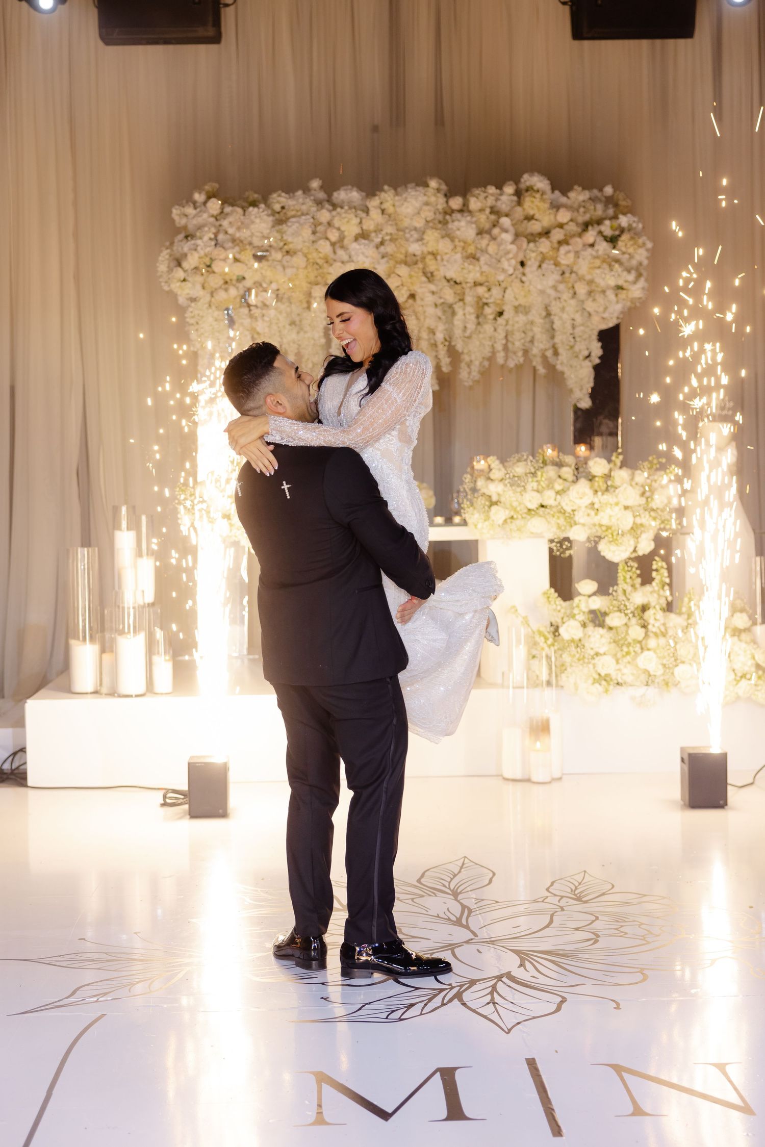 Bride and groom embrace during wedding first dance with white floral wall and sparklers in background.