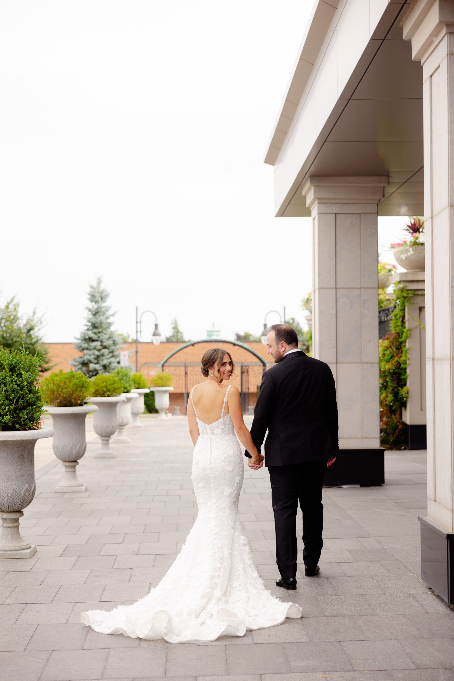 A bride and groom walk hand in hand along an elegant columned porch with decorative urns and stone pavers.