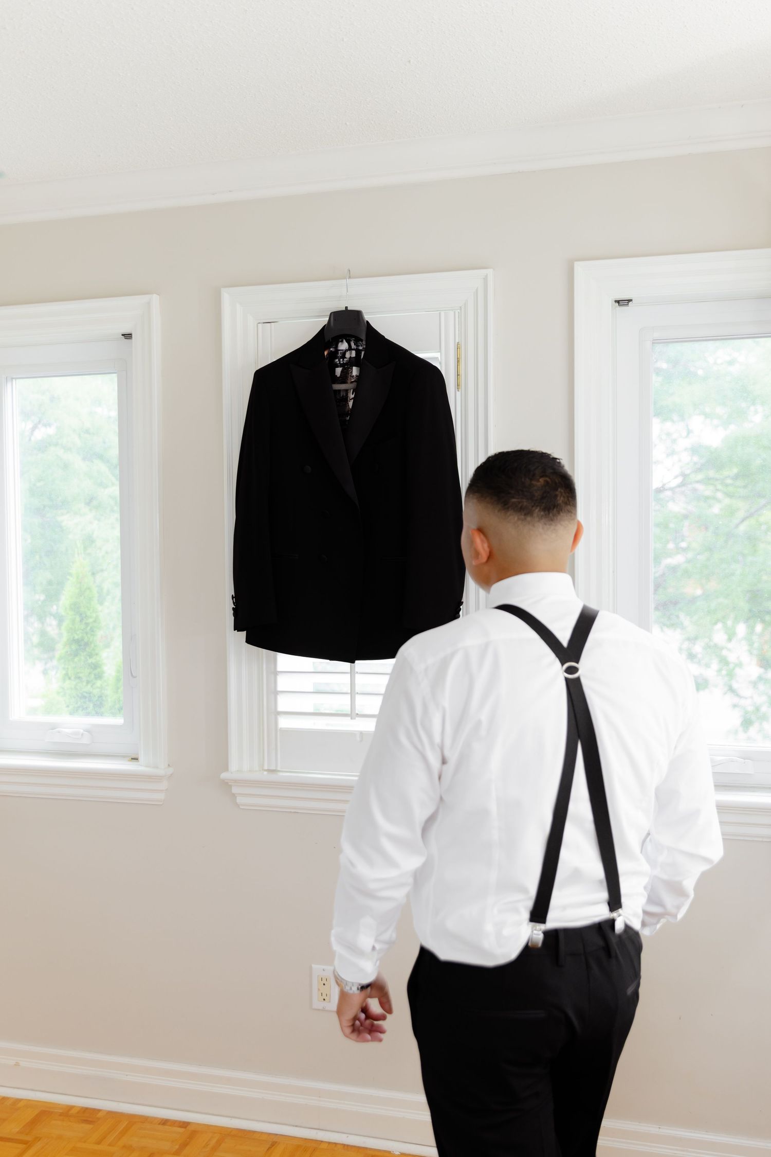 A person in formal attire with suspenders and a white dress shirt looks out a bright window in a minimalist room.
