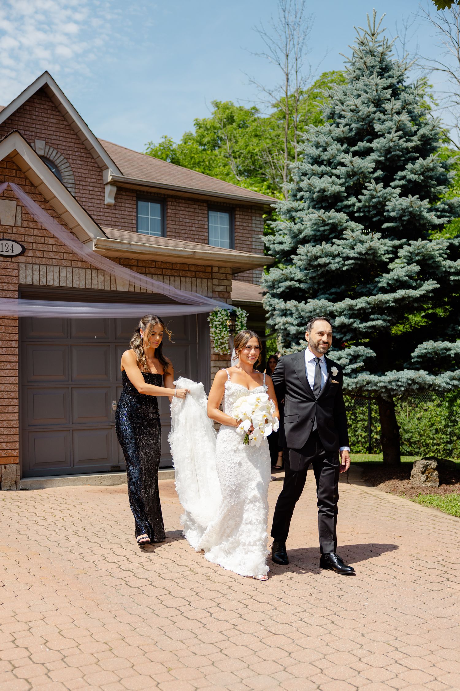 Two people walk on a paved driveway beside a brick house with a tall blue spruce tree on a sunny day.