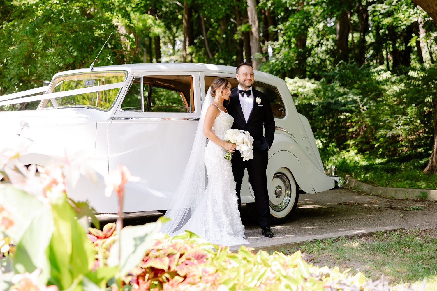 A wedding couple stands beside a classic white vintage car in a lush garden setting.