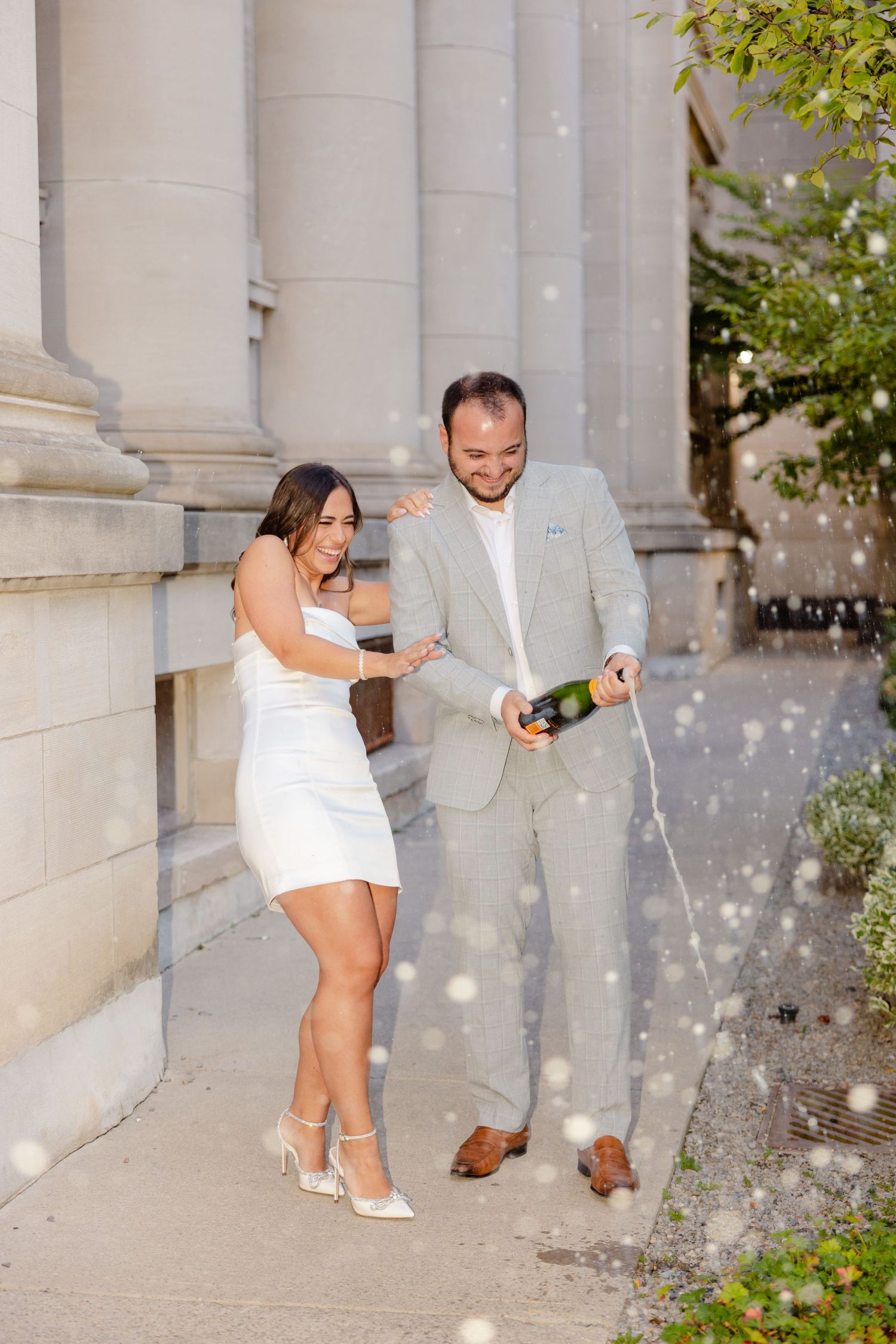 Couple enjoying champagne spray moment against white columns.