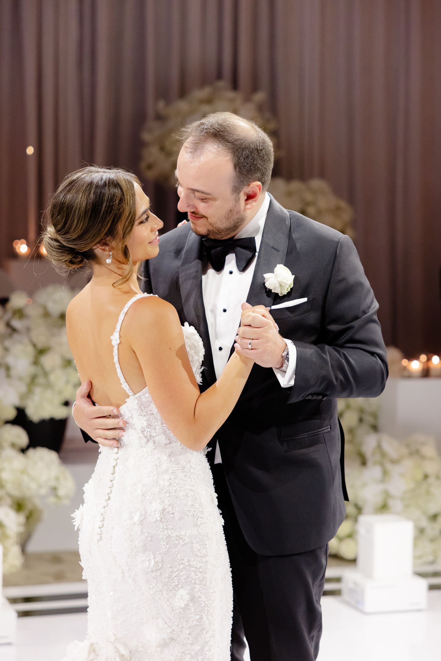 Newlyweds share their first dance at a wedding reception with white floral decorations and brown curtains in background.