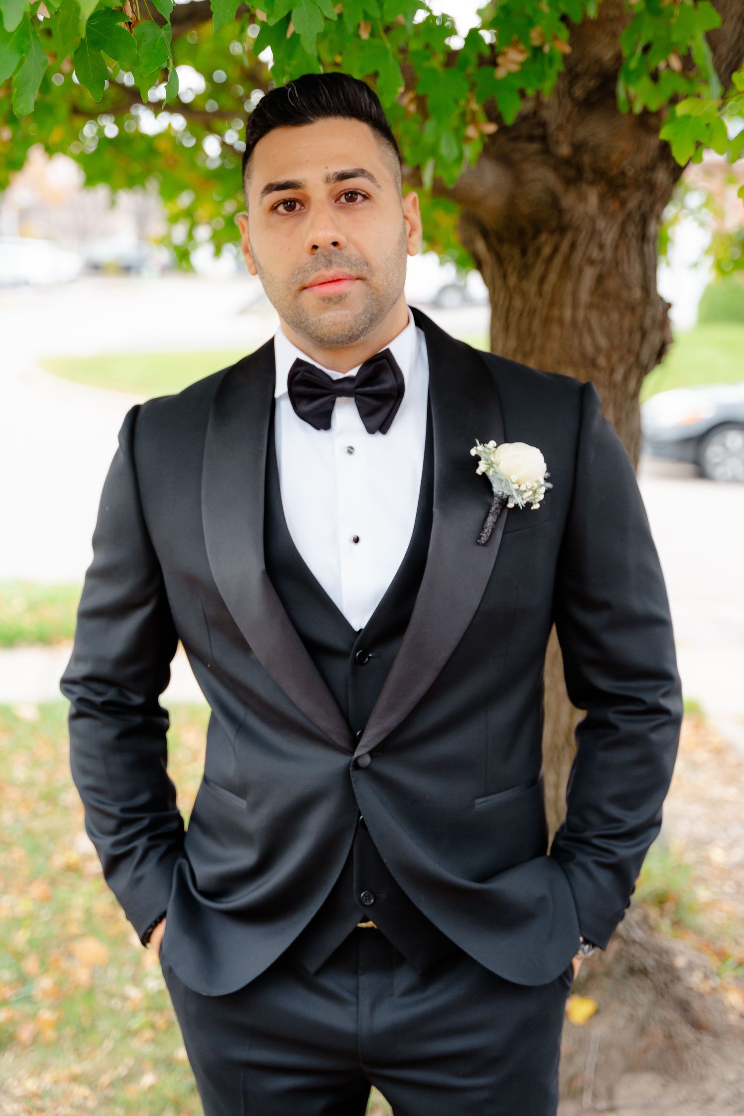 Elegant groom in three-piece tuxedo with white boutonniere poses under green leaves.