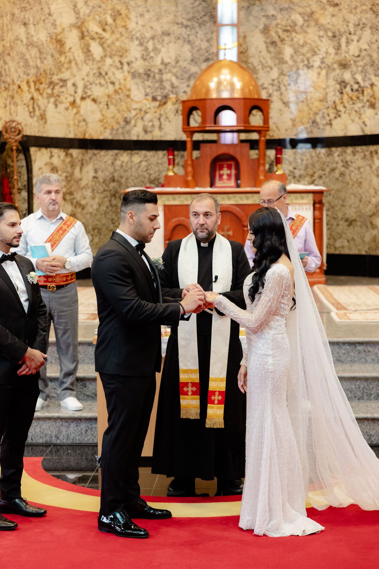 A wedding ceremony taking place at an ornate church altar with red carpet and gold walls.