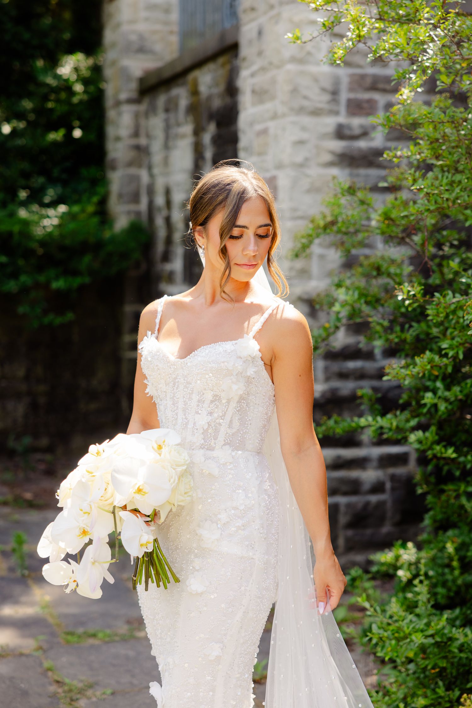 A bride in a white lace wedding dress and veil holds a bouquet of white orchids against a stone wall background.