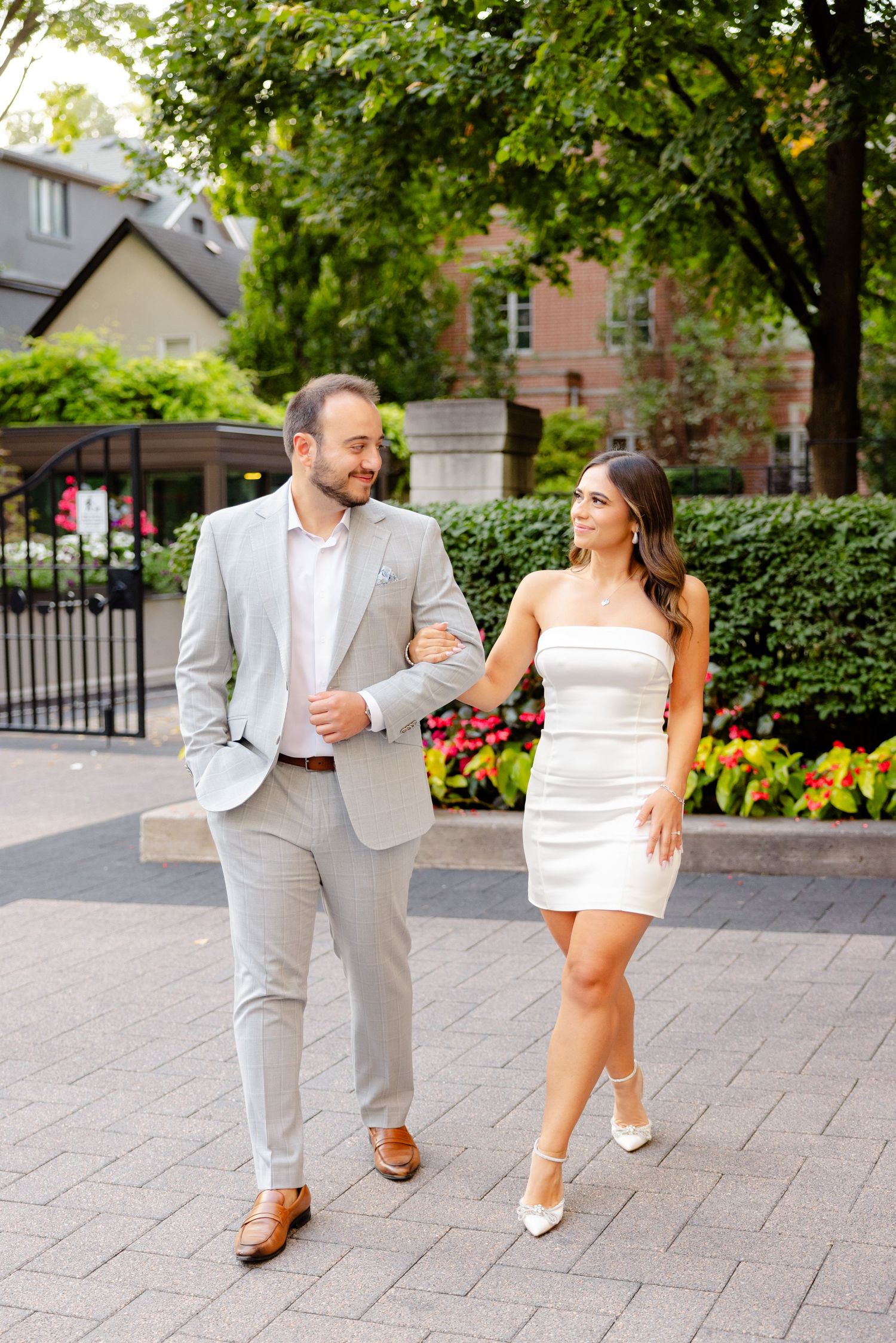 Stylish couple walking together on paved path with lush garden and decorative gate in background.