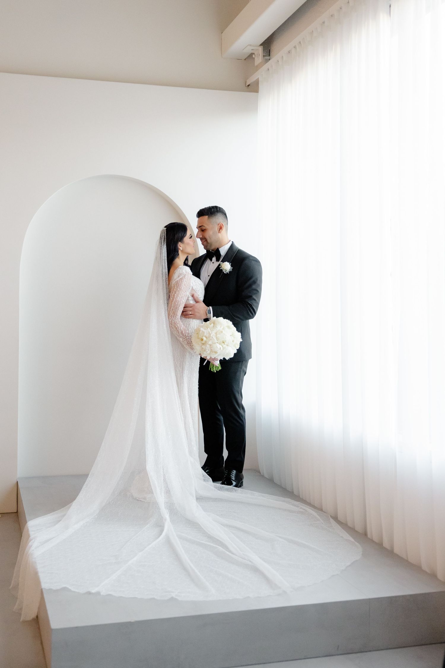 A wedding couple shares an intimate moment under an arched doorway with flowing white veil.