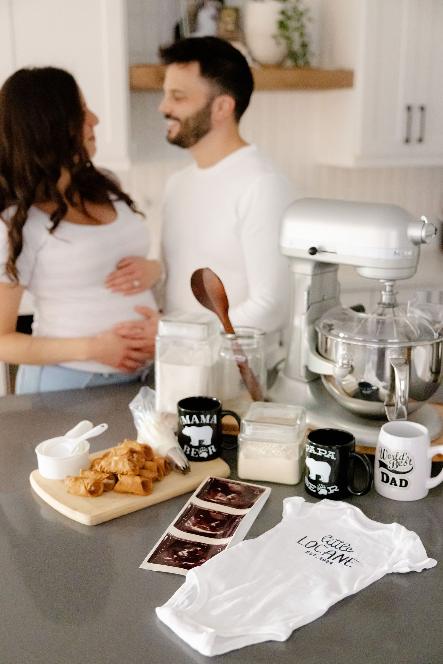 Couple in kitchen with baking supplies and baby items hinting at pregnancy announcement.