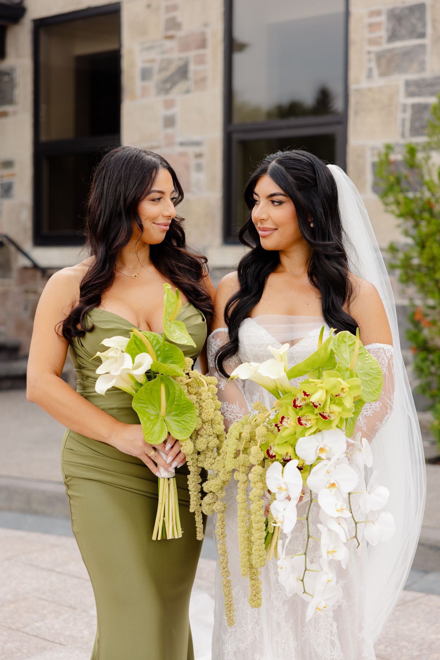 Bridesmaids in olive green dresses share joyful moments with the bride holding elegant white and green floral bouquets.