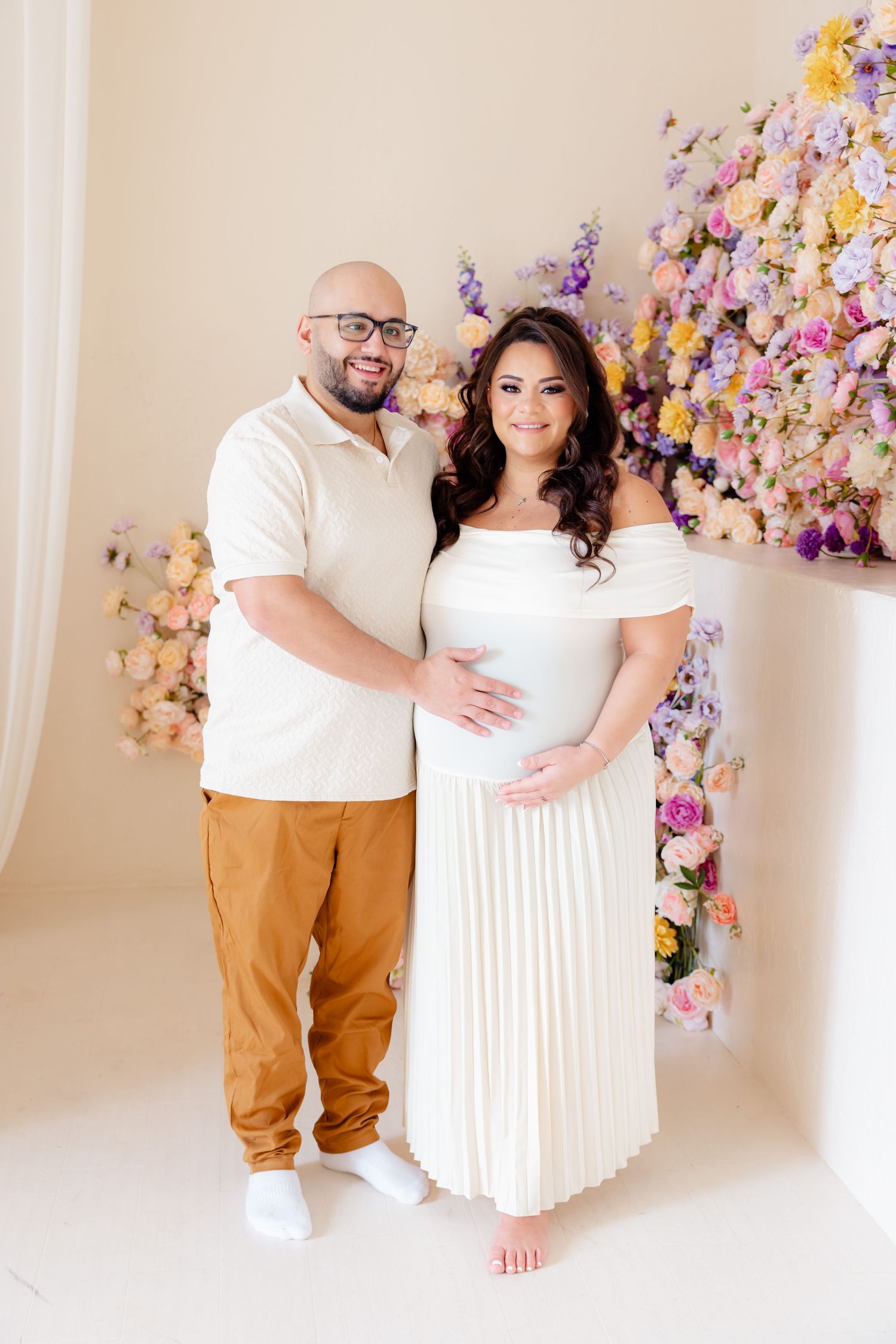 A couple poses in white outfits against a wall decorated with pink and purple floral arrangements.