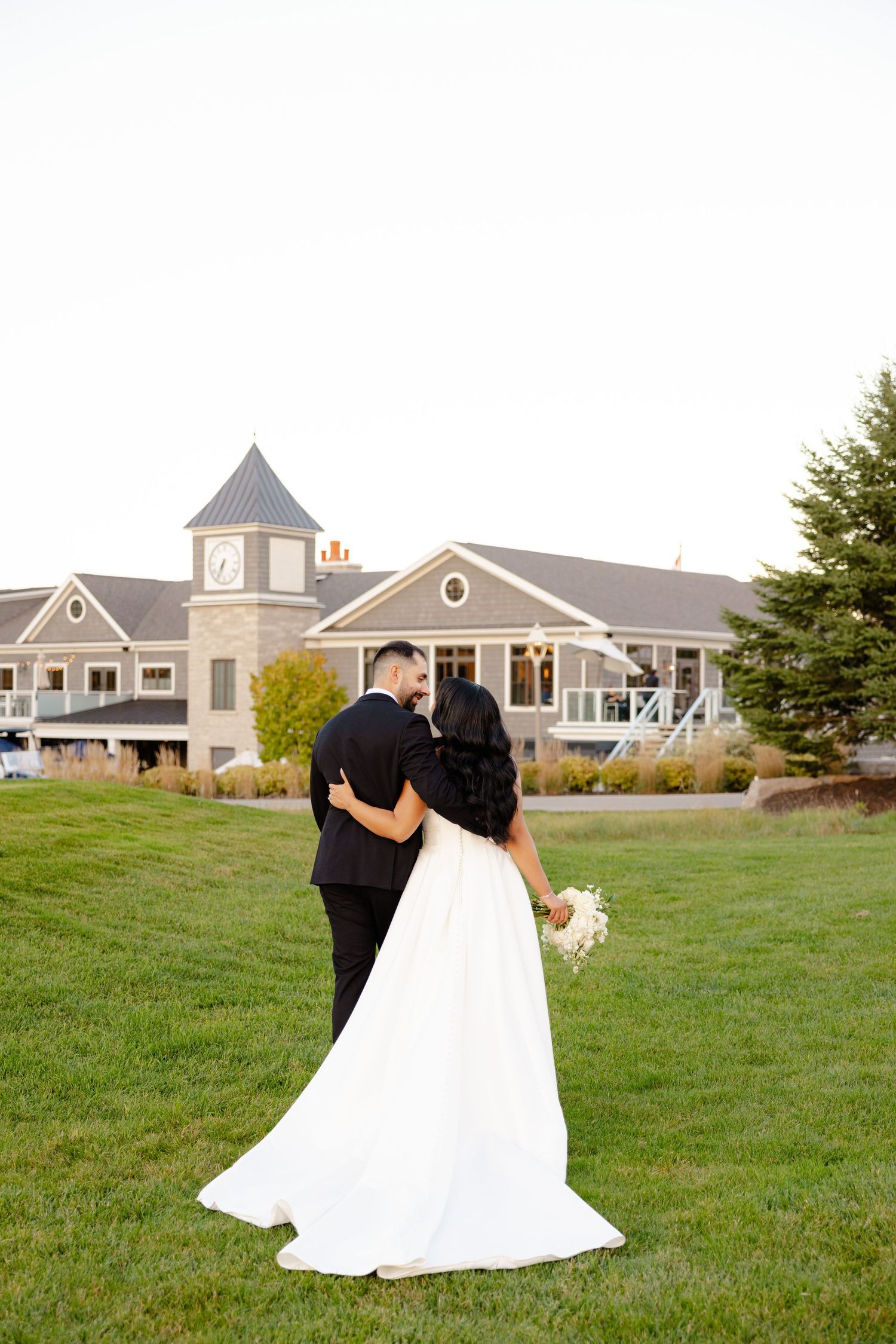 A wedding couple embraces while standing on manicured grounds with a large clubhouse in the background.