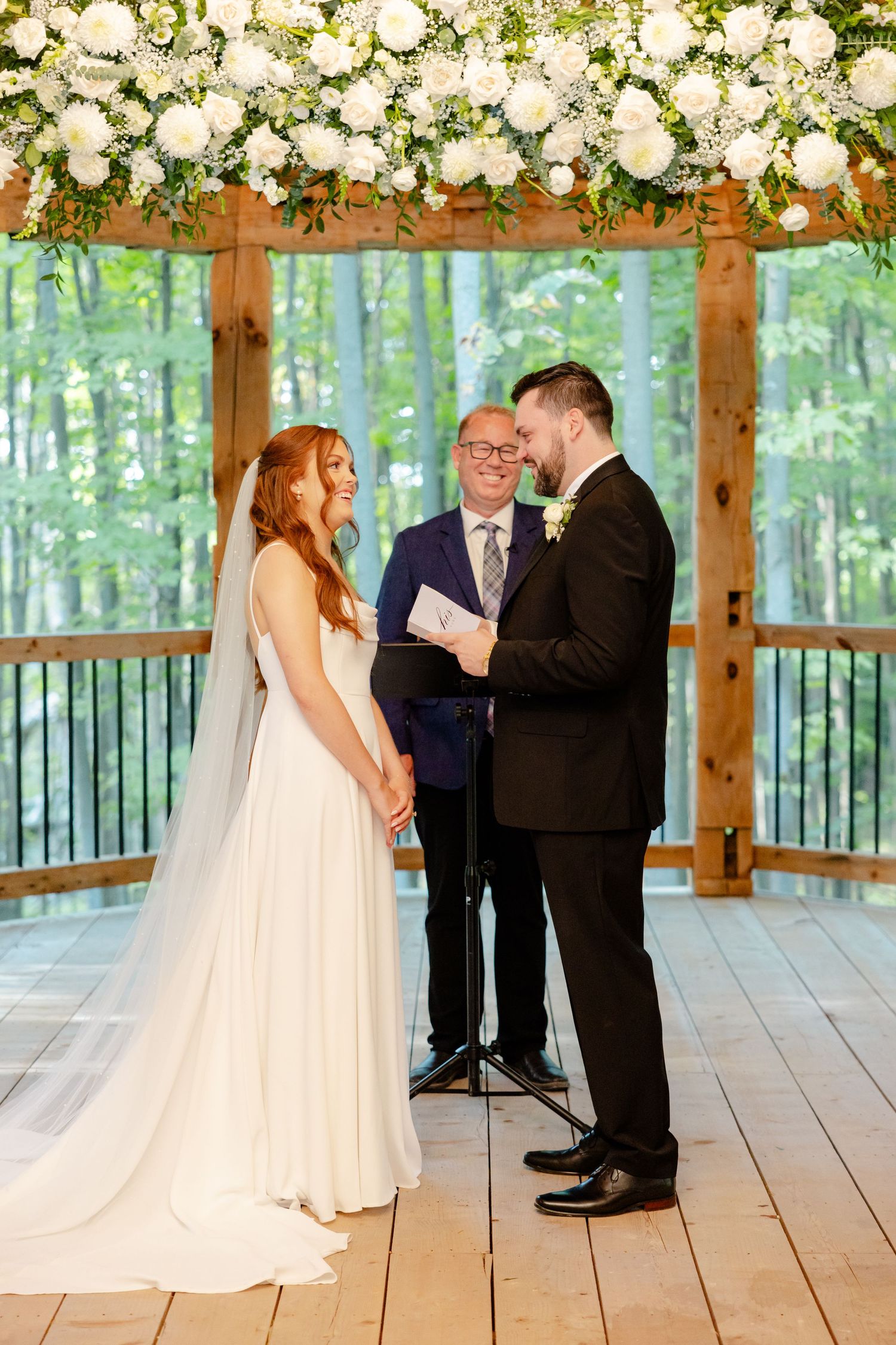 Wedding couple exchanging vows beneath white floral arch in forest pavilion setting.