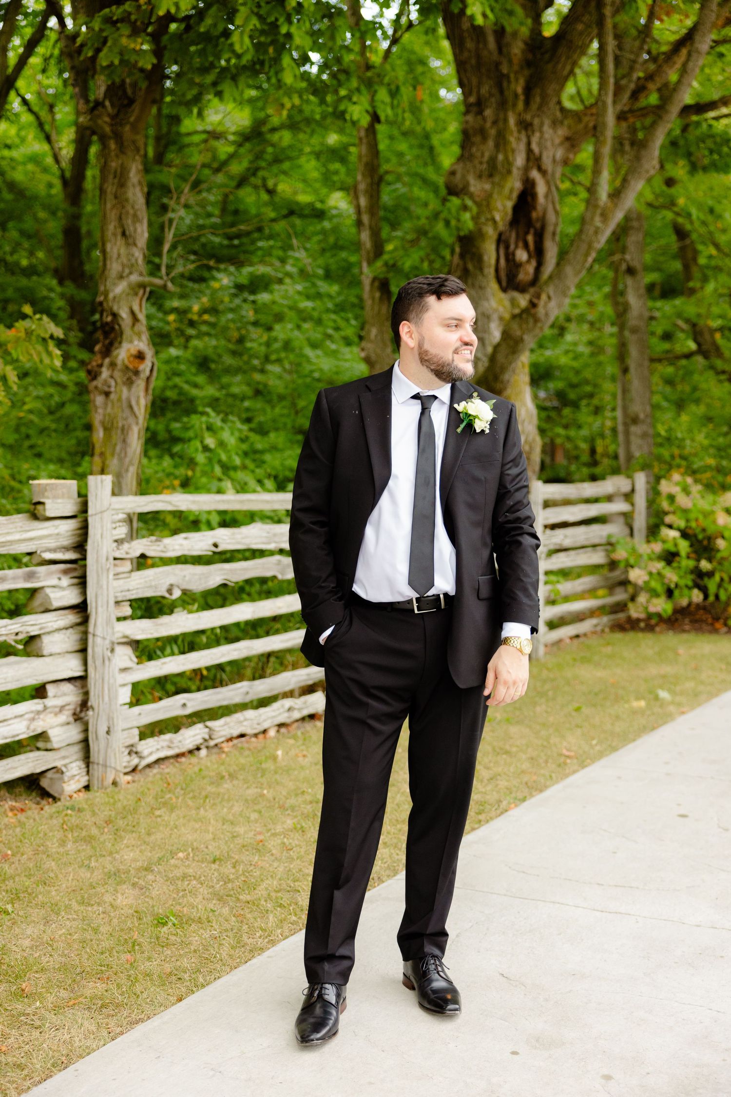 Groom in formal black suit poses casually by wooden fence and pathway.