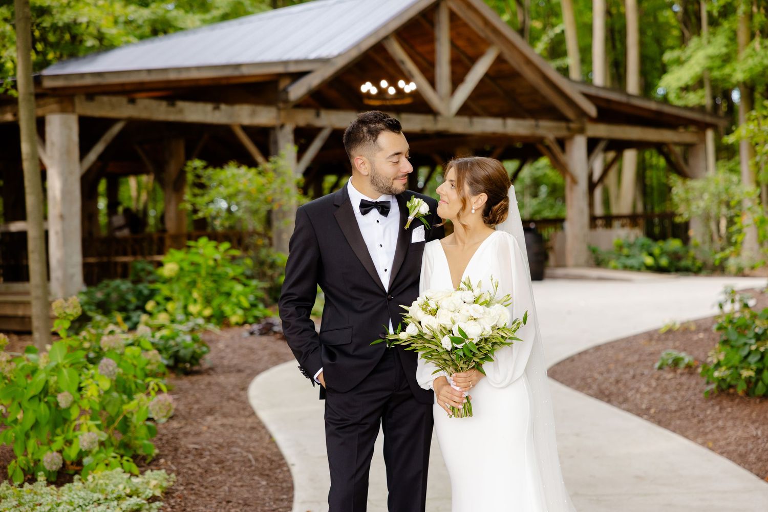 Elegant couple shares romantic moment on winding path with woodland pavilion setting.