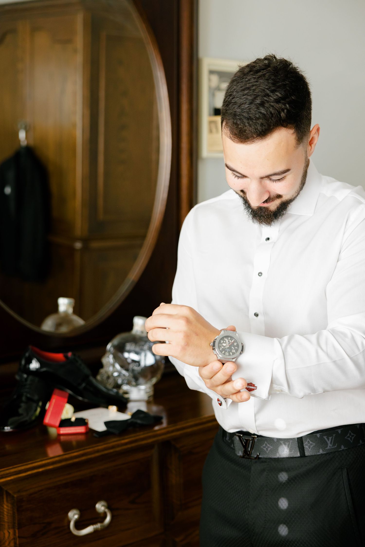 Person adjusts a silver watch while wearing a crisp white dress shirt in front of a wooden dresser.