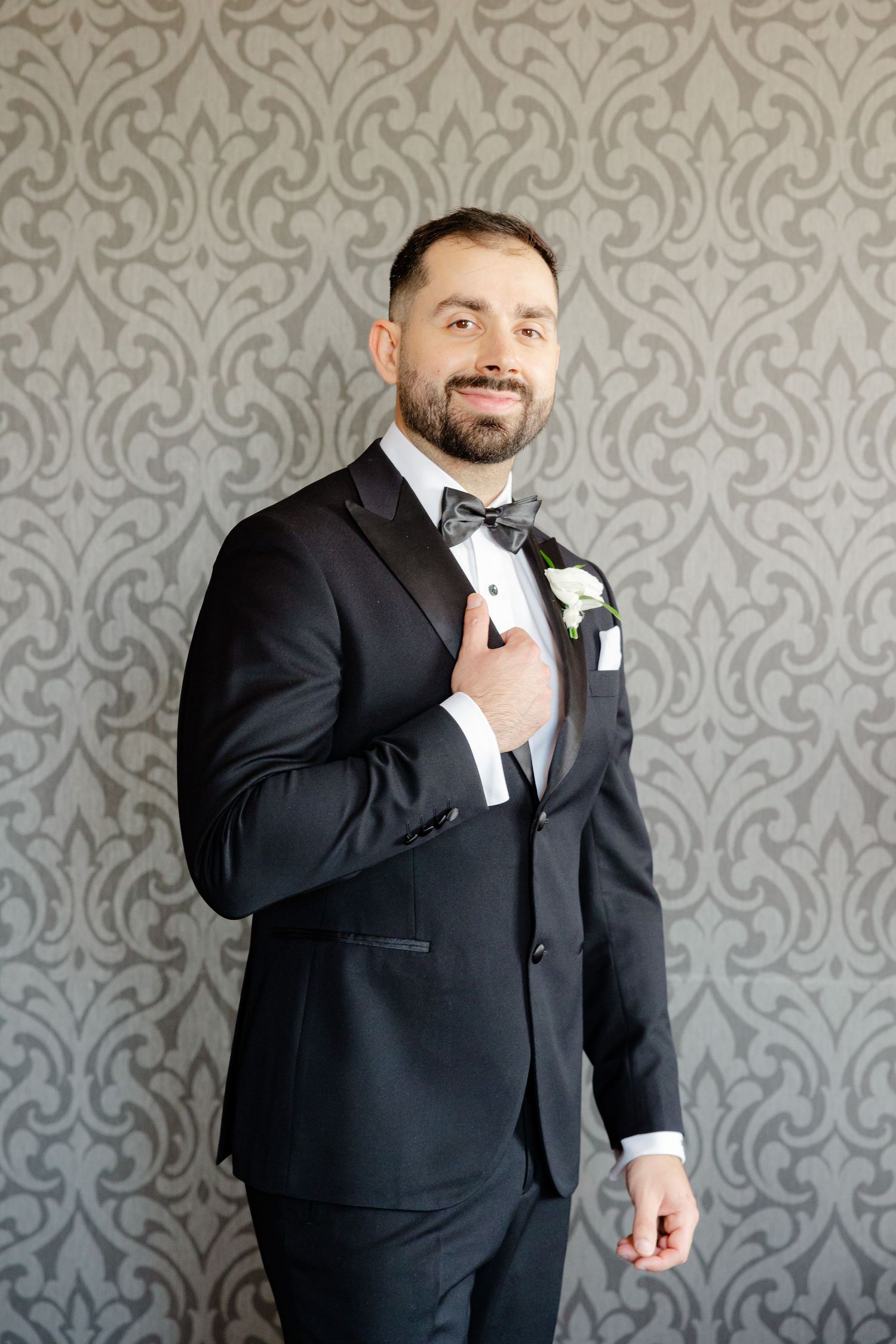 A formal portrait of someone in a black tuxedo with bowtie against a damask wallpaper background.