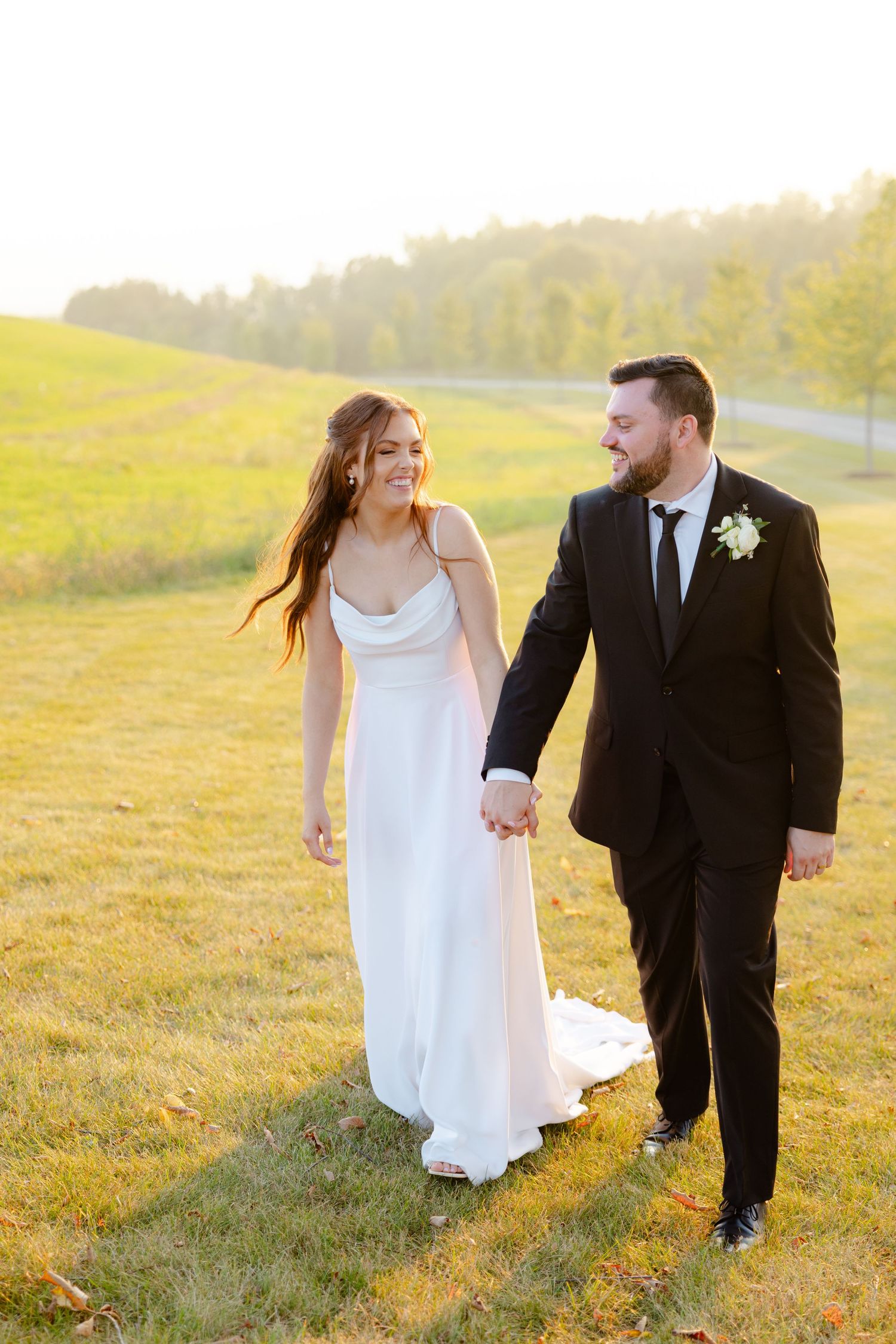 Joyful newlyweds walk hand in hand through a sunlit field with wind-blown hair.