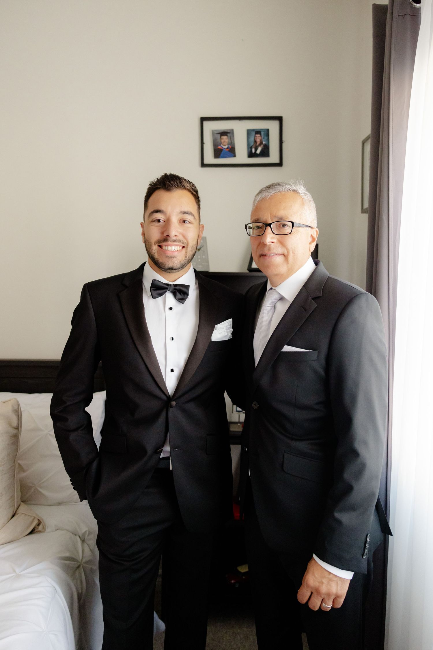 Two people in formal attire smile together in bedroom setting.