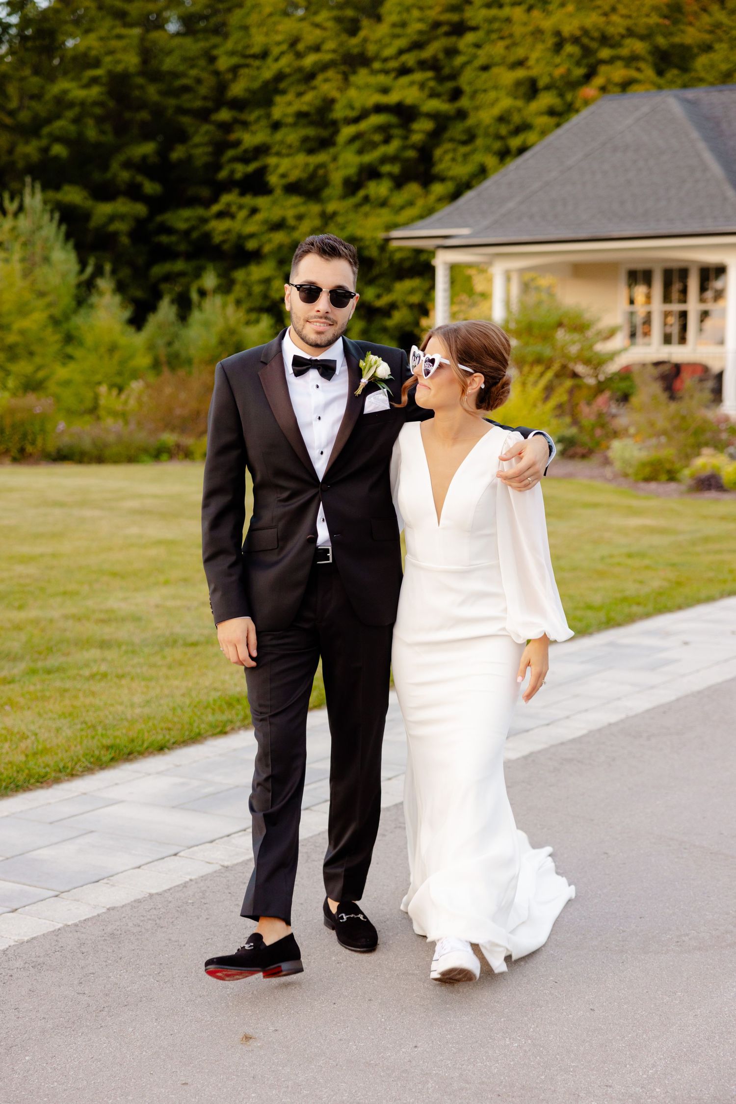 A stylish wedding couple wearing sunglasses walks together on a paved pathway.