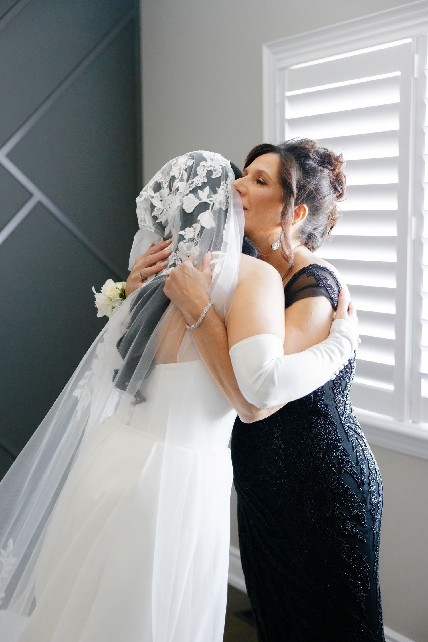 An emotional moment shared between a bride and a woman in a navy dress near white shutters.