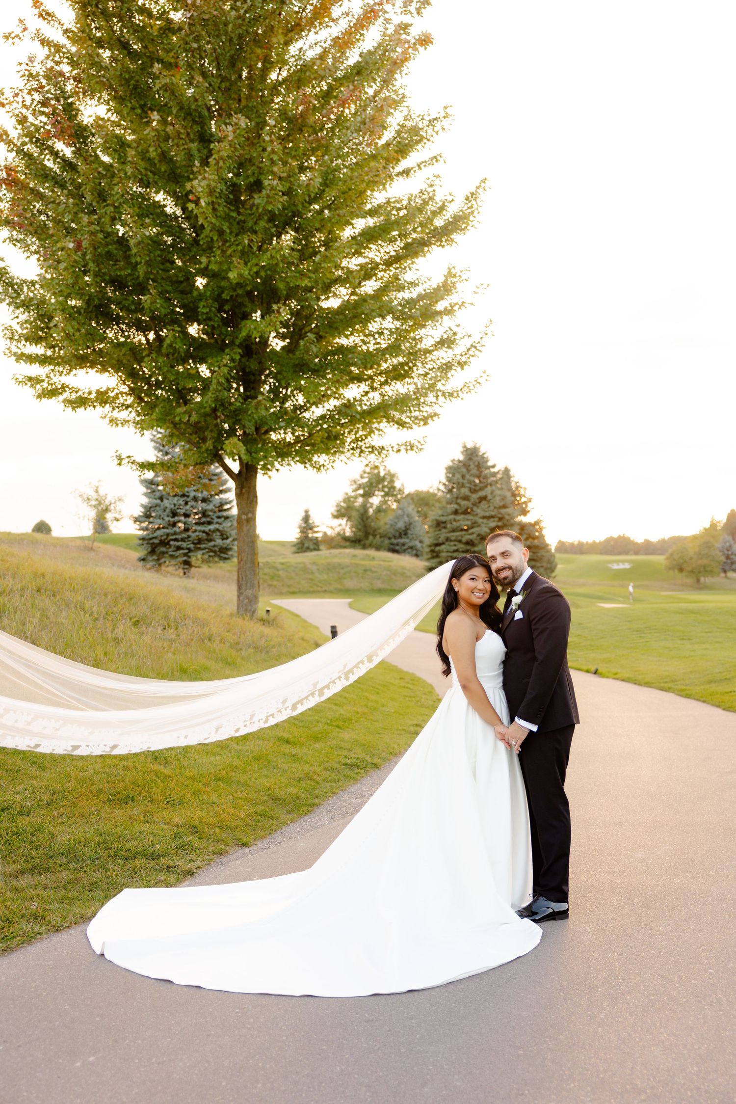Wedding couple standing on a path with a dramatic flowing veil under a large tree.