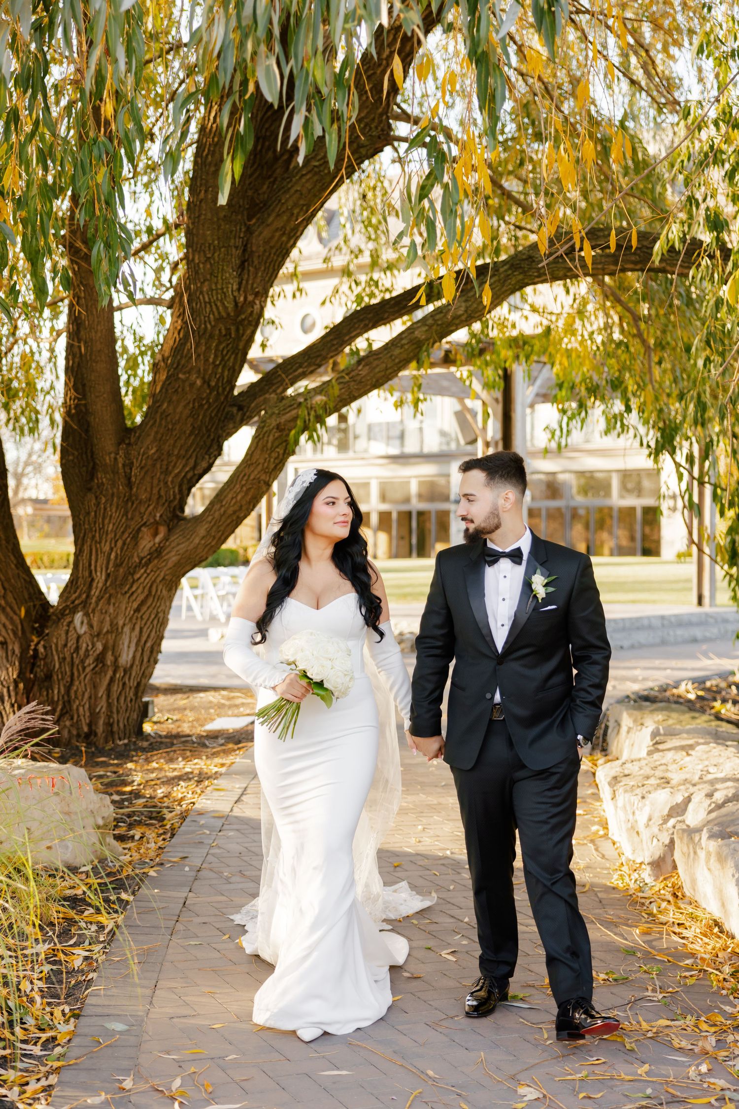 Elegant wedding couple walking together on a fall day under drooping willow branches.