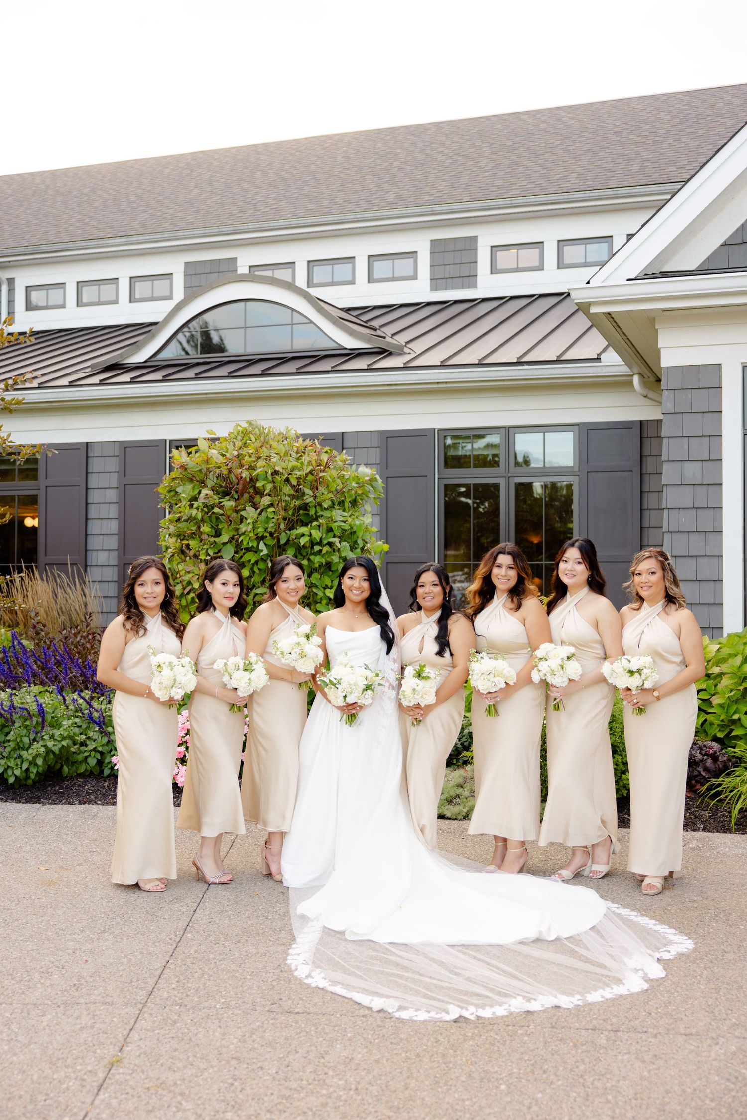 A bridal party in cream-colored dresses poses in front of a modern gray building with landscaped gardens.
