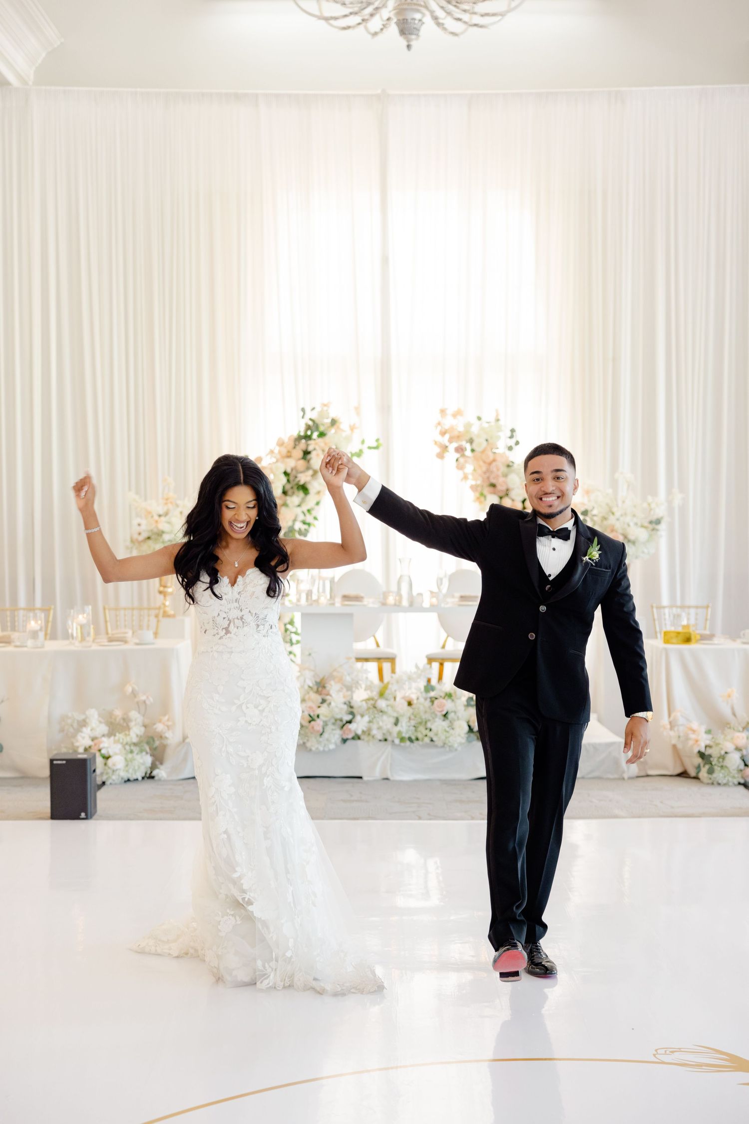 Joyful wedding celebration dance in white ballroom with floral decor and chandelier lighting.