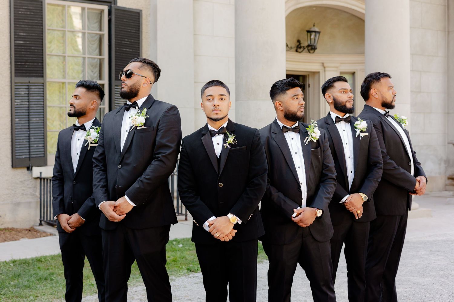 Groomsmen line up in black tuxedos for a formal portrait against a classic building facade.
