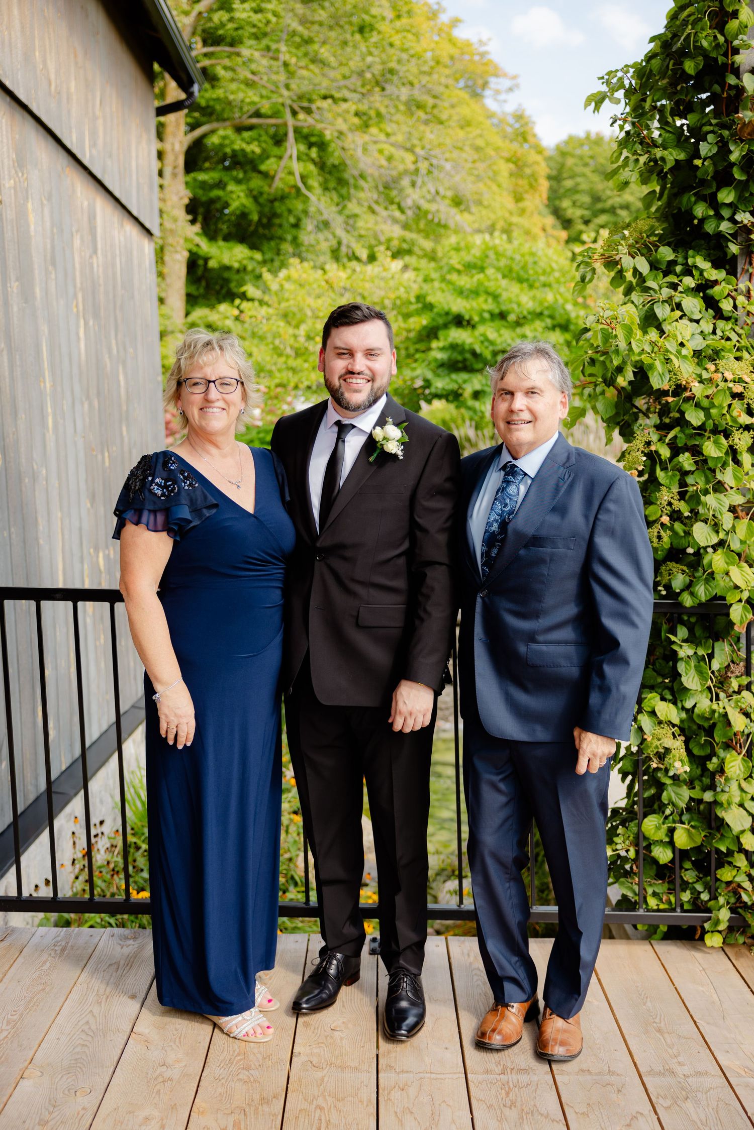 Family poses together on wooden deck with lush greenery background at formal event.