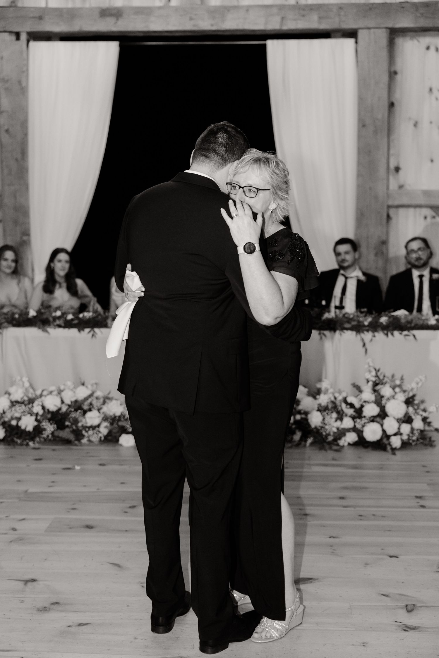 A black and white photo captures an emotional dance moment at a barn wedding reception.