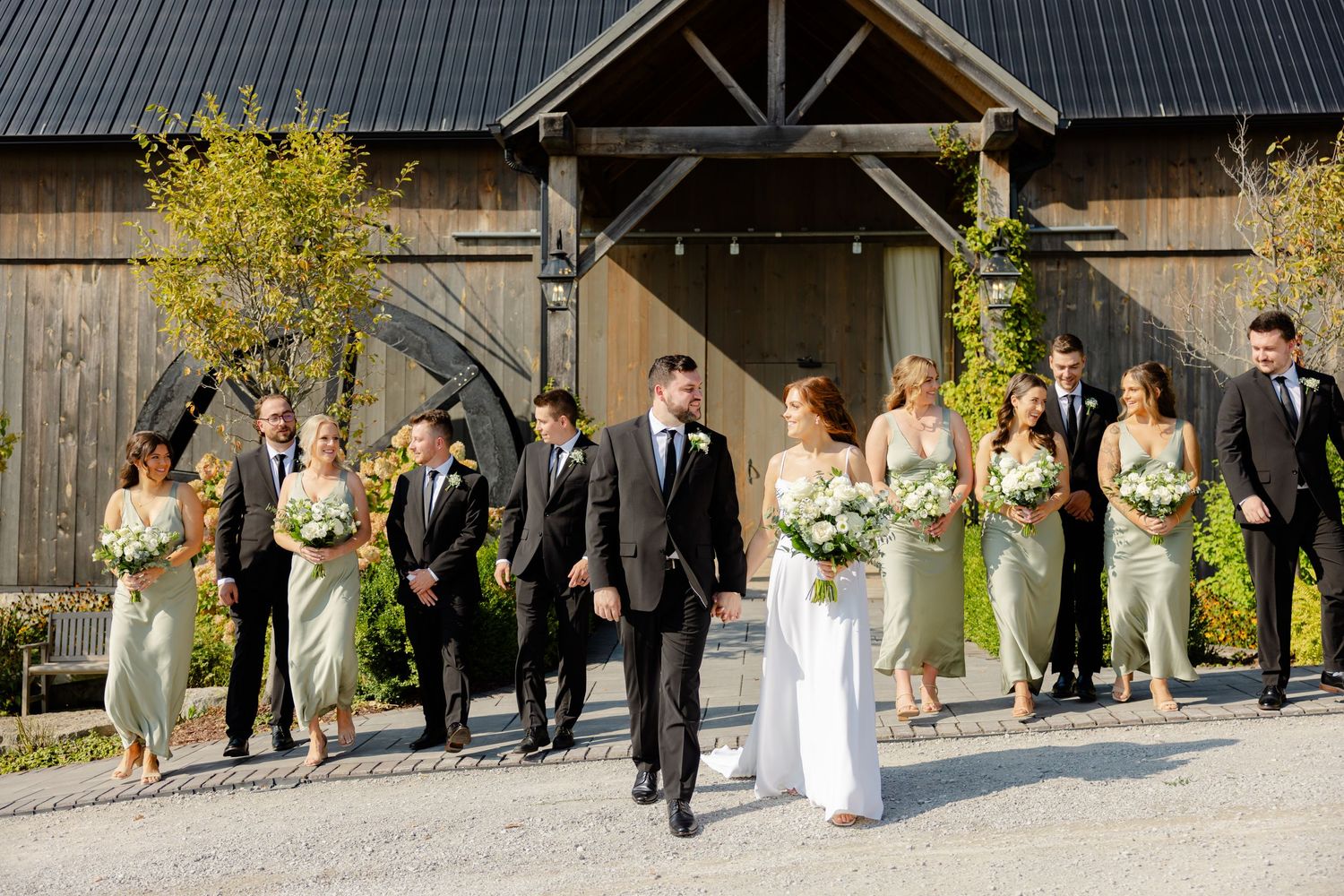 Wedding party walks together outside rustic barn venue wearing sage green and black attire.