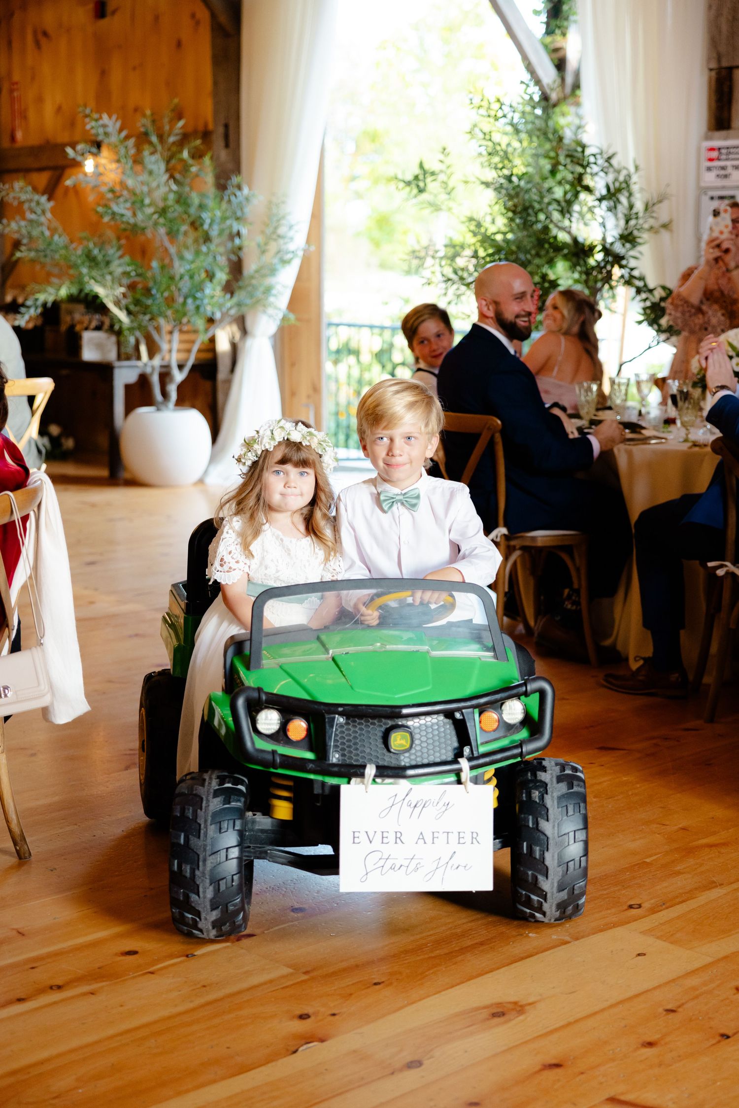 Two young attendants ride in a green miniature vehicle during an elegant indoor wedding reception.