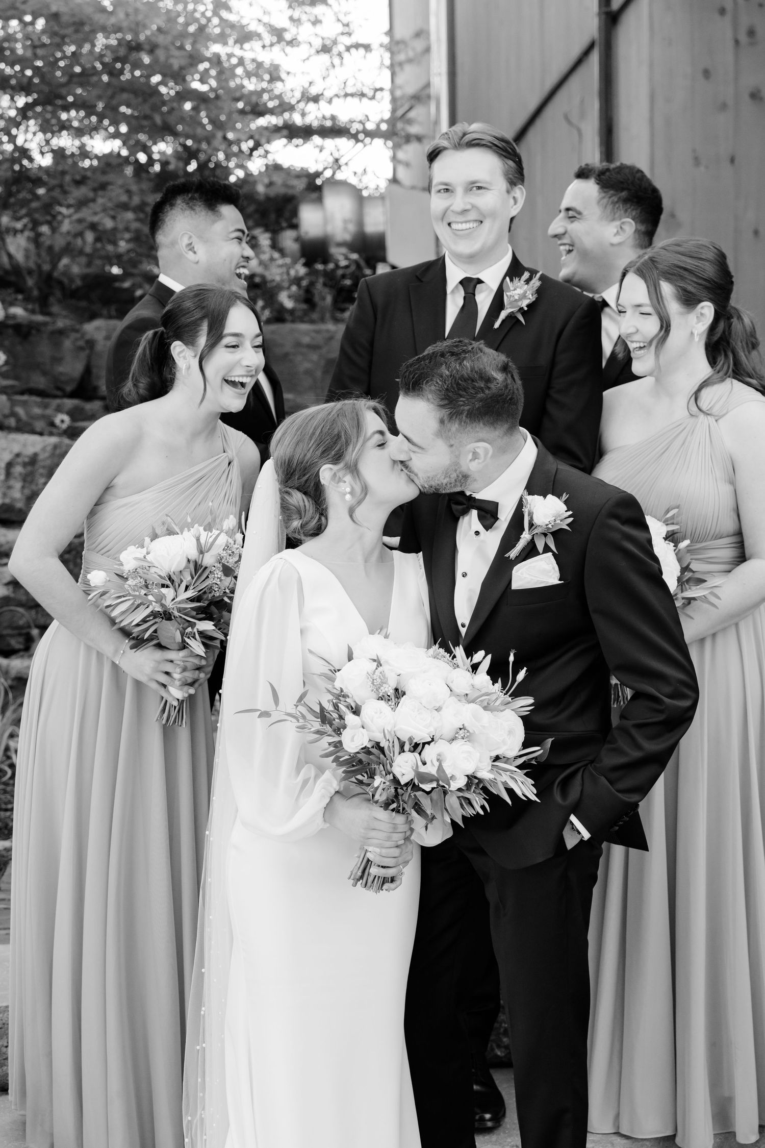 Black and white candid photo of wedding party sharing laughs during formal portraits.