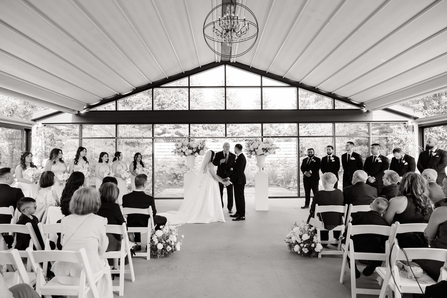 Black and white photo of indoor wedding ceremony with guests seated on white chairs.