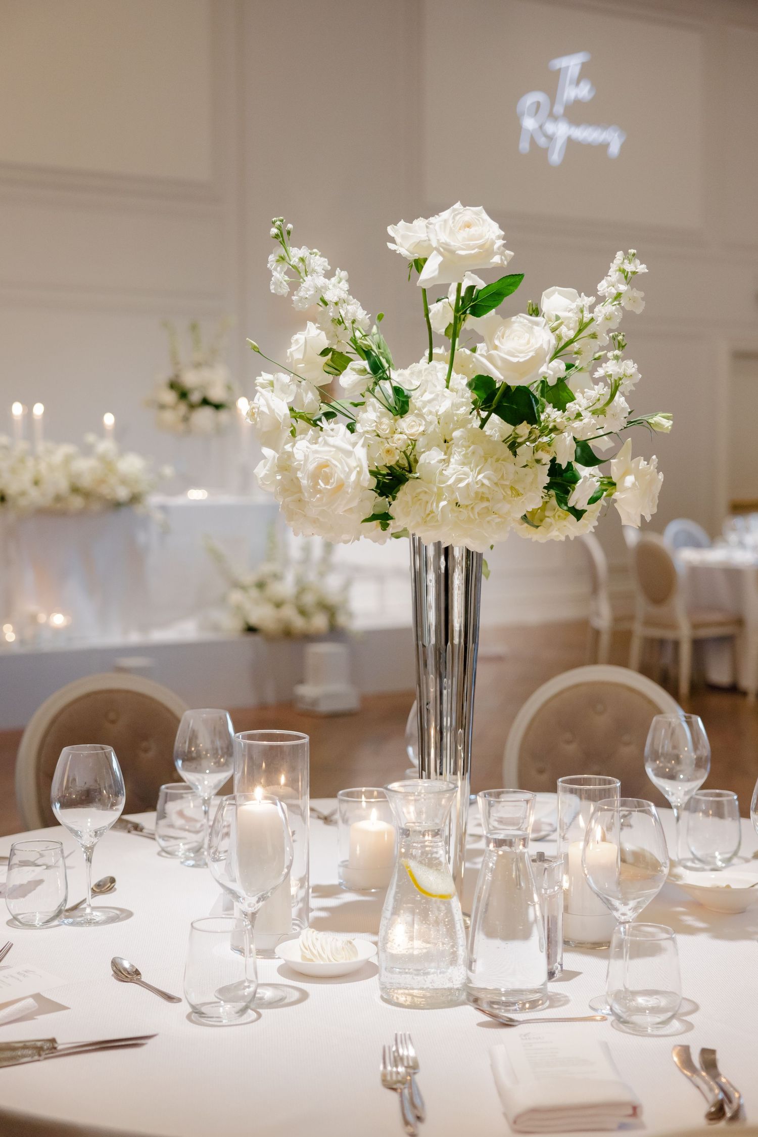White roses and delicate flowers in tall glass vase centerpiece with surrounding glassware on round table.