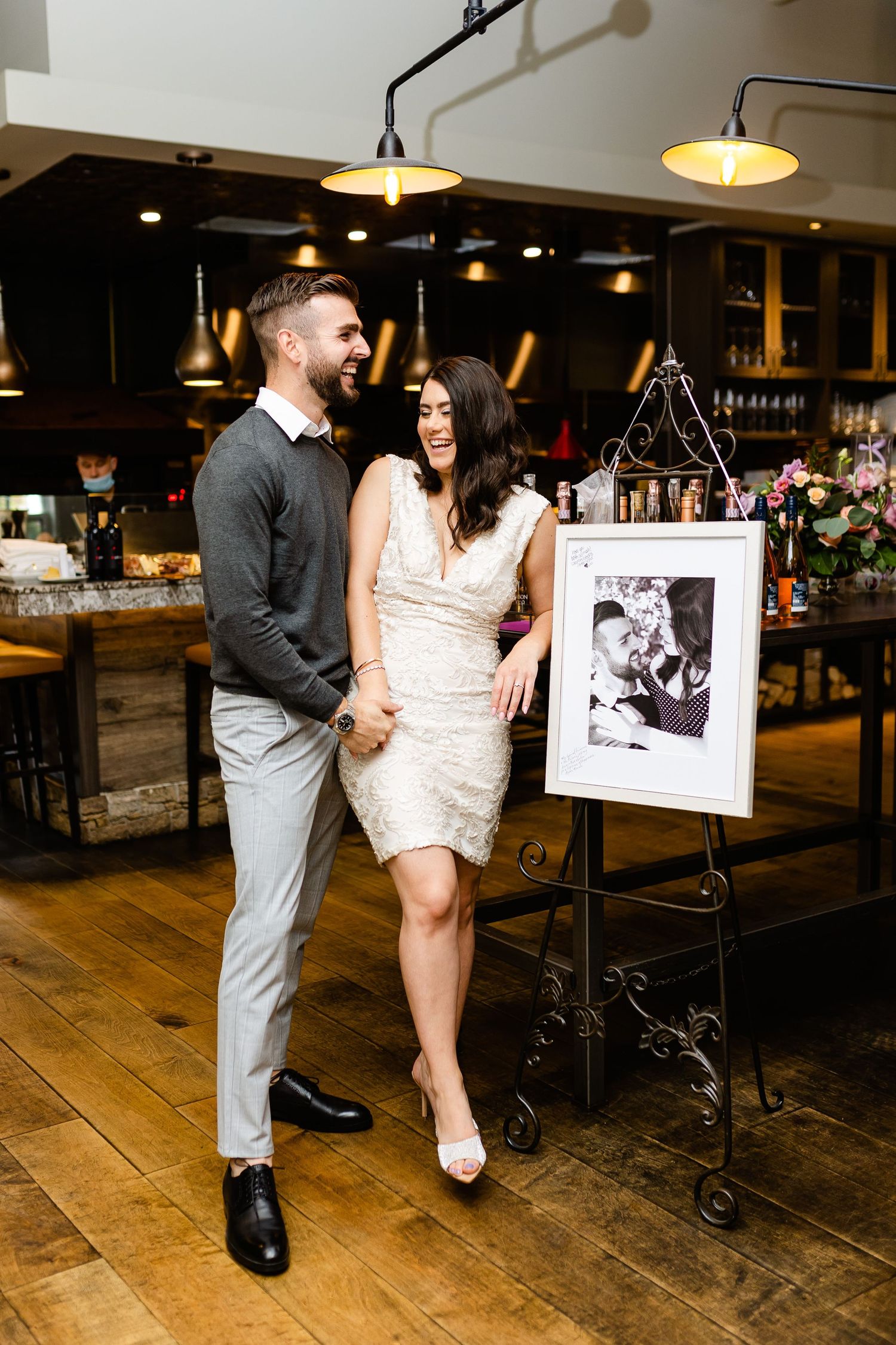 Laughing couple in formal wear standing by decorative easel in restaurant setting.