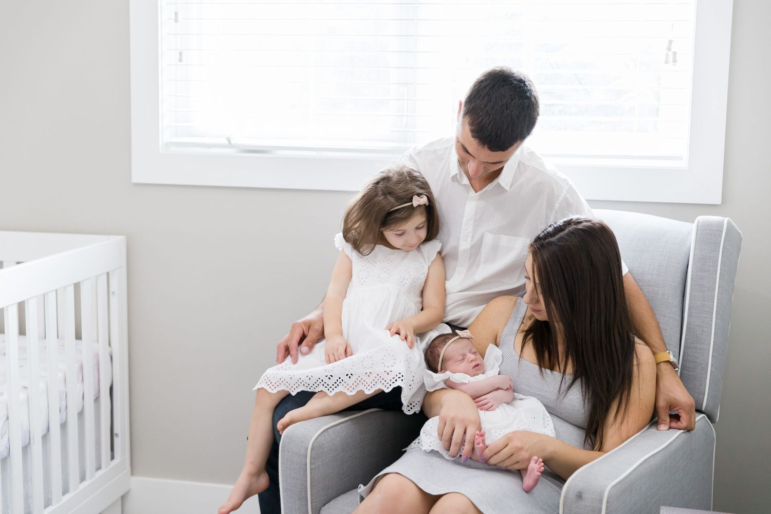 Family sitting together in a cozy grey armchair next to a white crib.