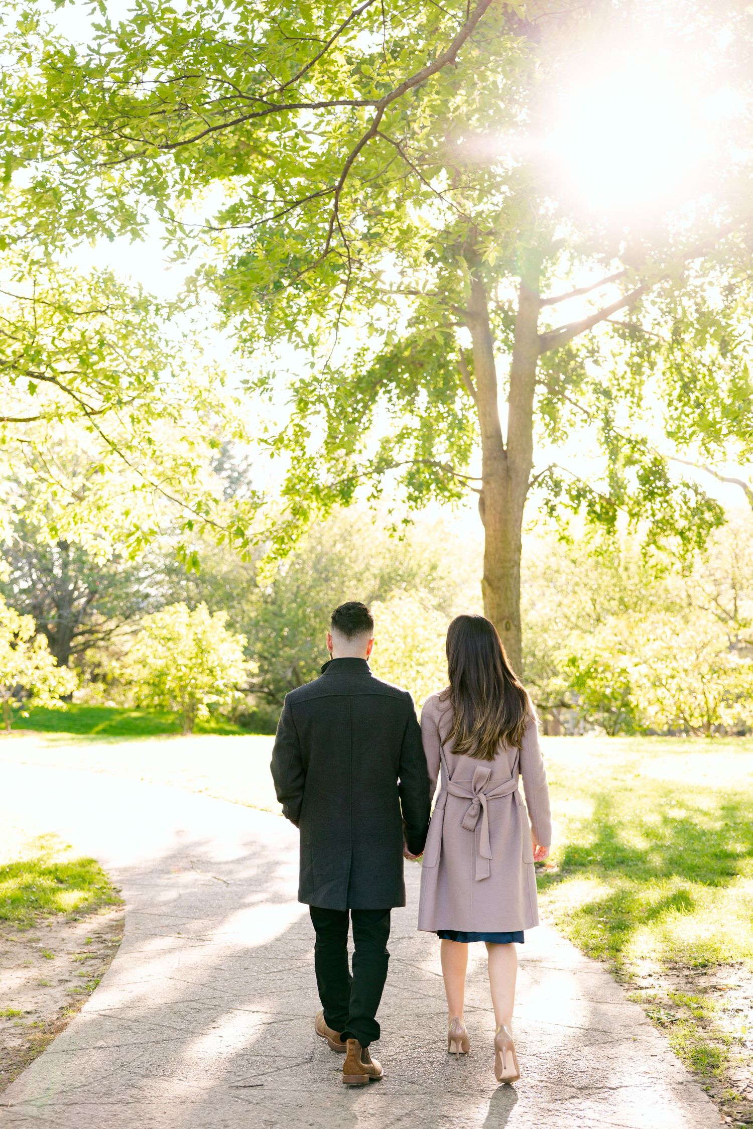 A couple walks hand in hand down a sunlit path beneath green spring foliage.