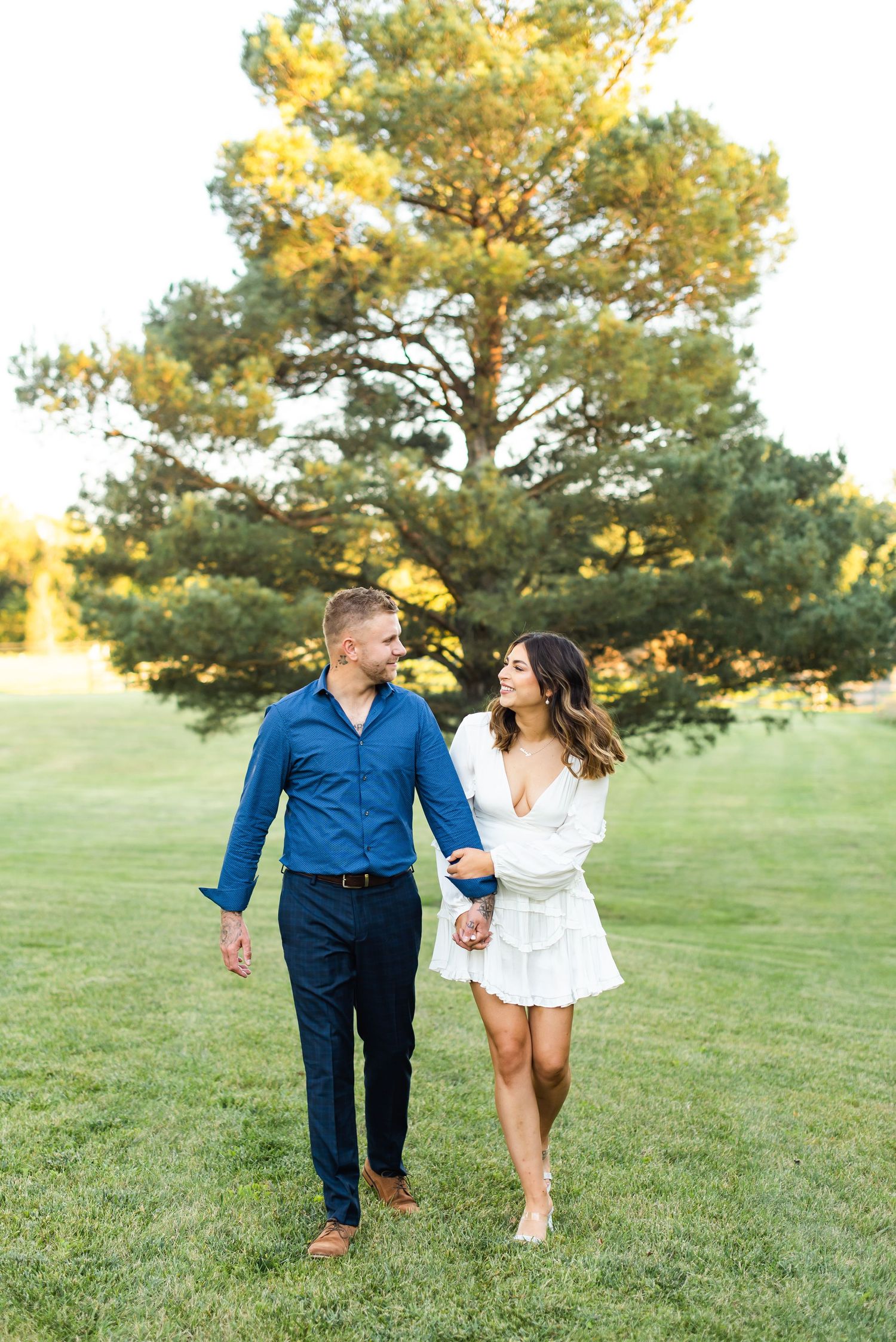 Happy couple taking romantic walk across grassy field with large pine tree behind them.