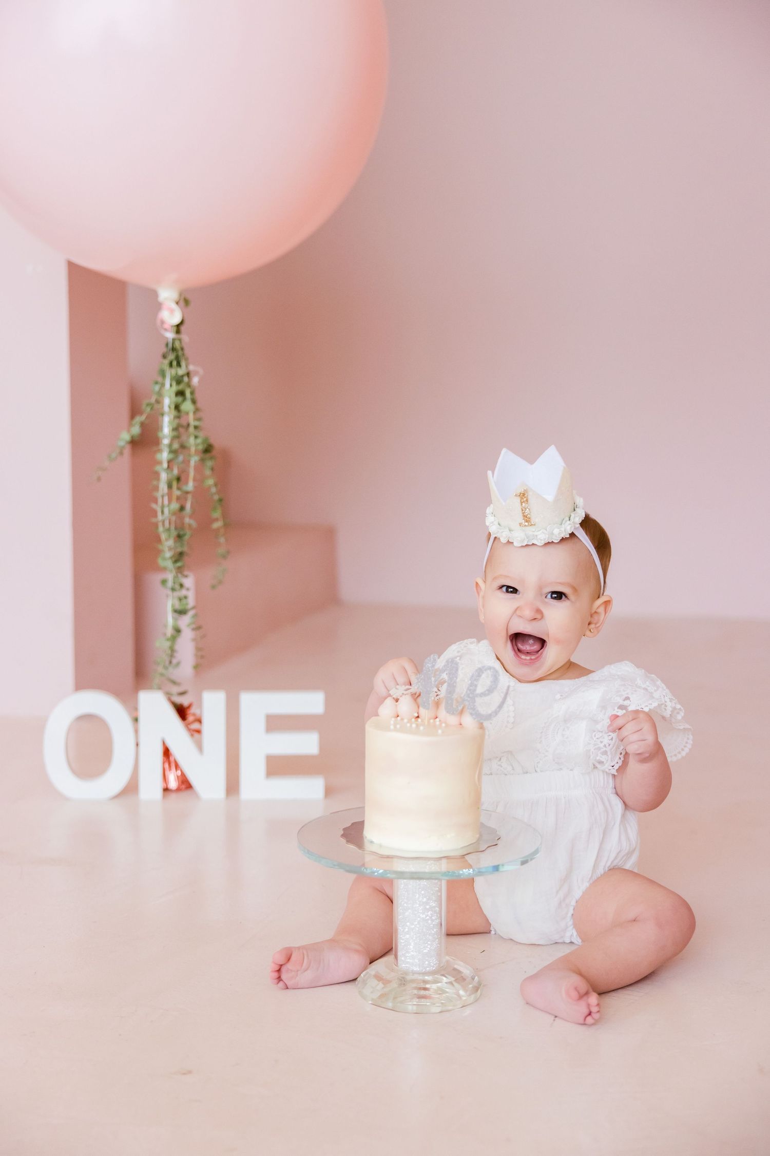Sweet first birthday cake smash photo with white cake, pink balloon, and white ONE letters against pink backdrop.