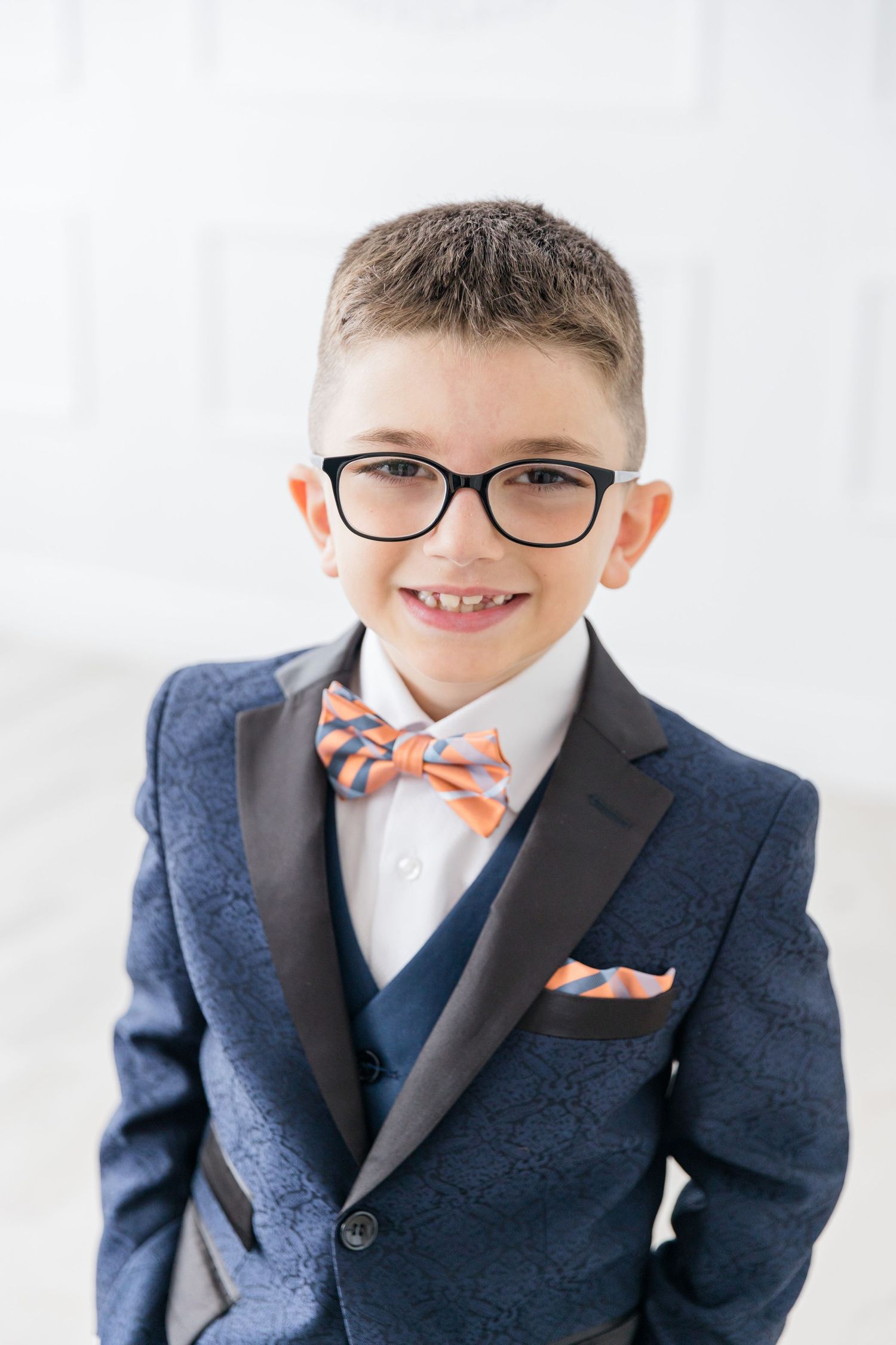 Young person wearing navy suit with glasses and orange polka dot bowtie smiles against white background.