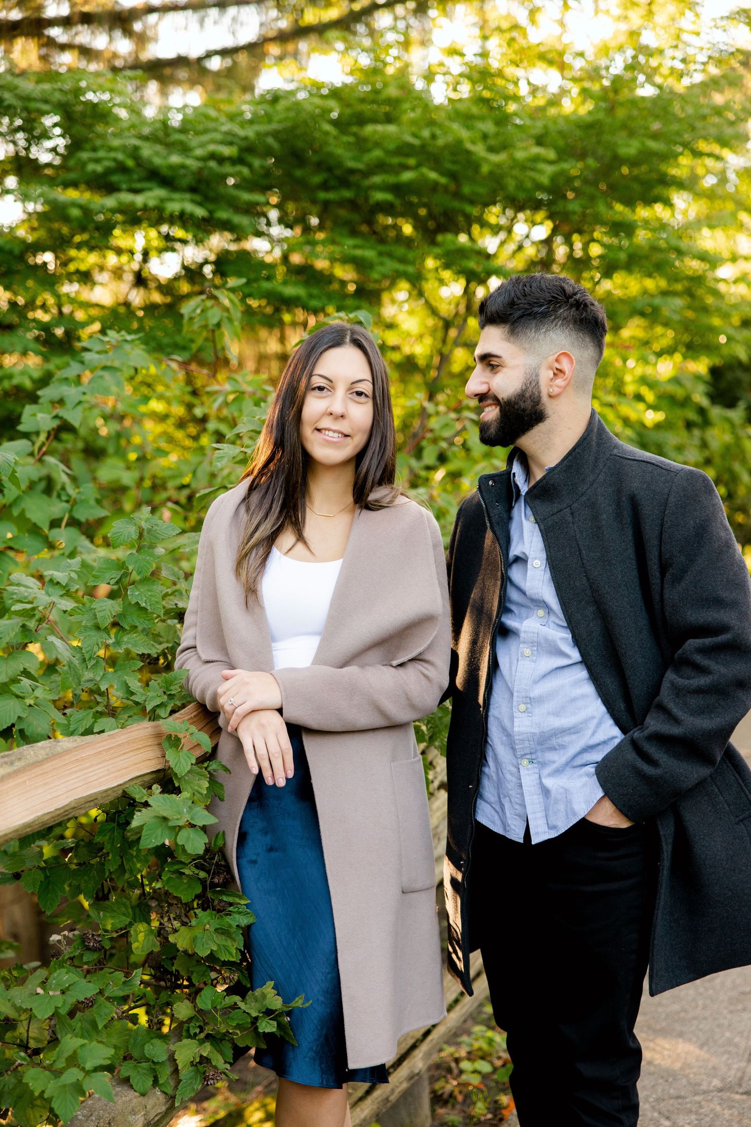 Couple in fashionable outerwear sharing a candid moment by wooden railing in park.