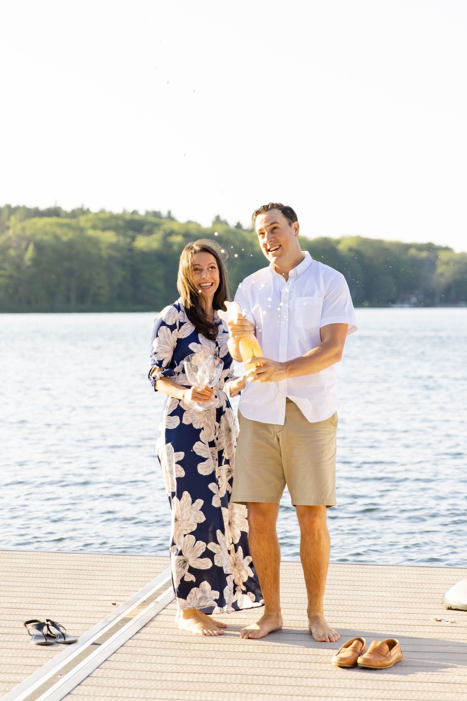 Couple in summer attire sharing drinks on sunny lakeside dock.