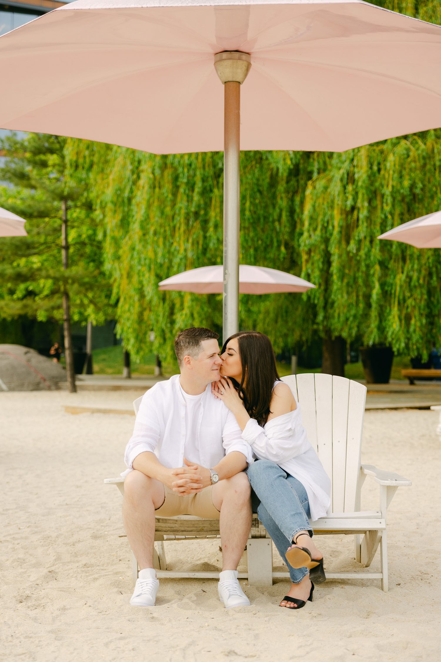 Romantic couple sharing kiss on beach chair under pink umbrella.